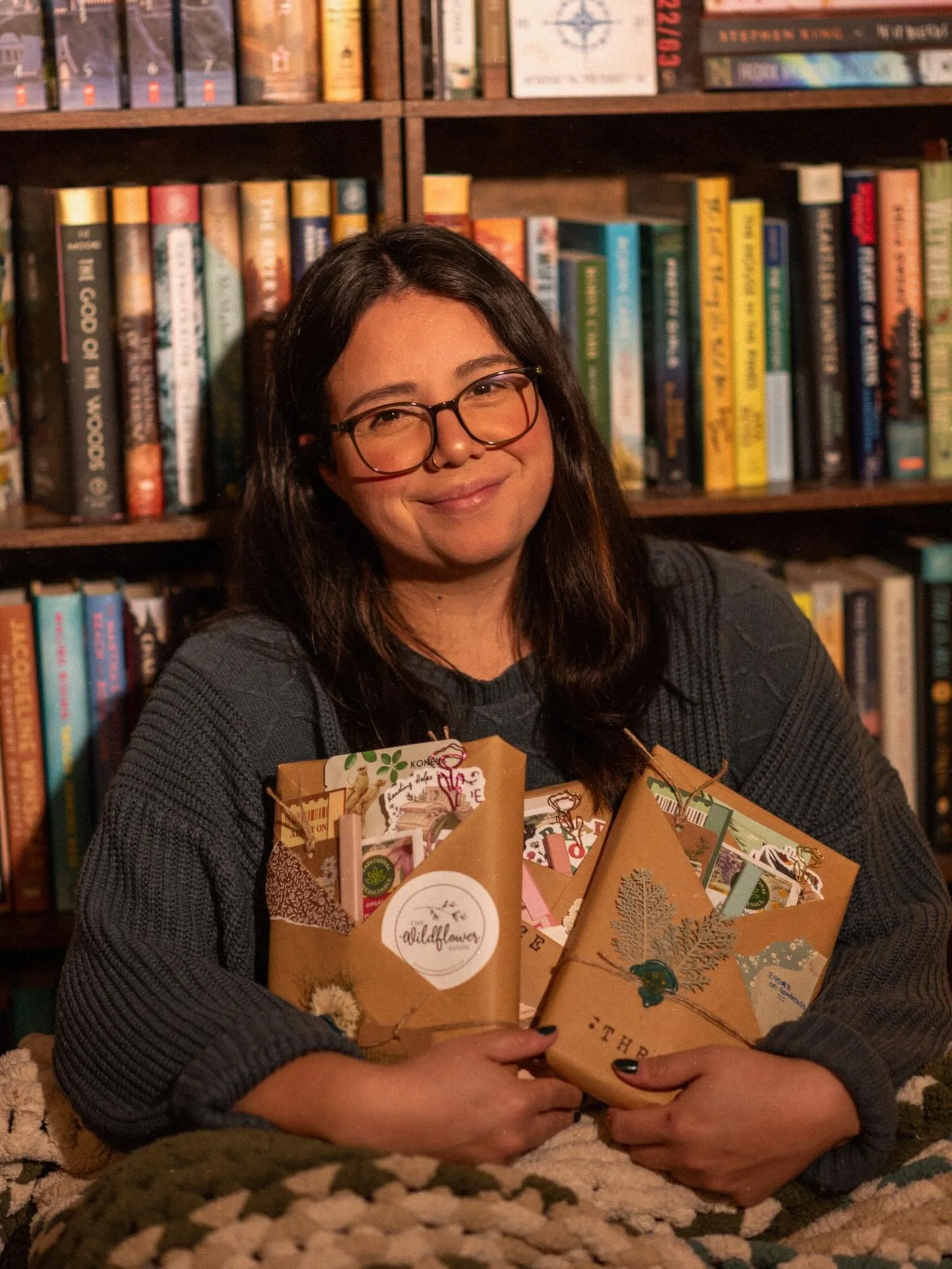 Woman with glasses smiling, holding holiday gift packages in a cozy room with a bookshelf in the background.
