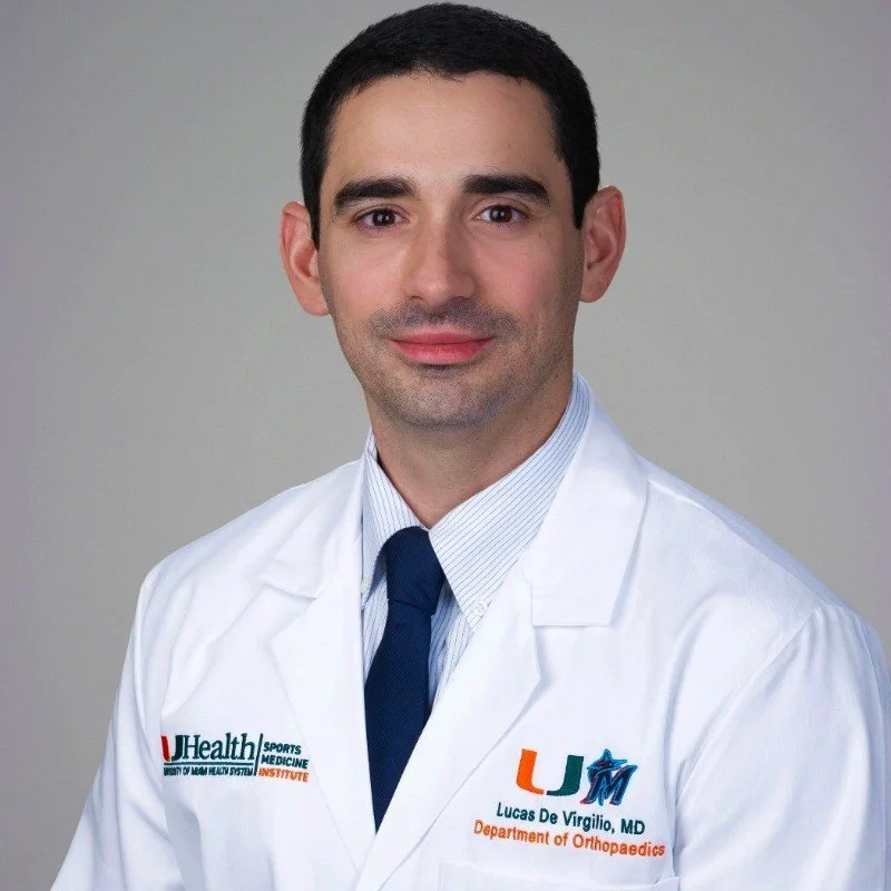 Professional portrait of a male doctor wearing a white lab coat with an embroidered logo, a dress shirt, and a tie, against a plain gray background.