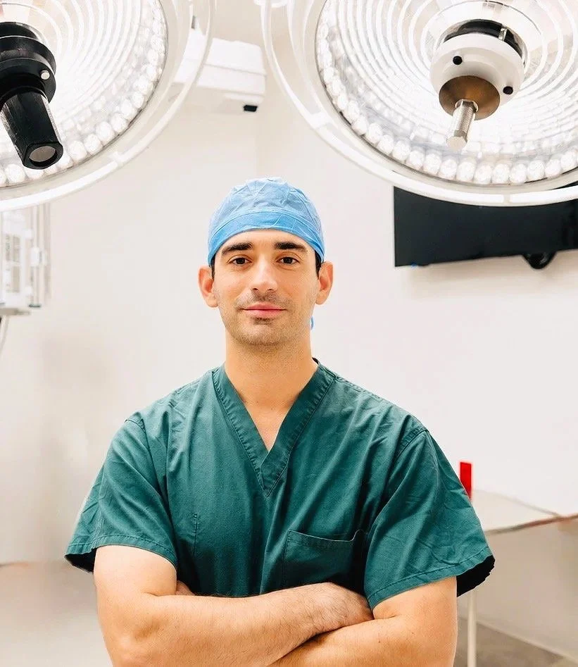 Male surgeon in green scrubs and blue surgical cap standing confidently in an operating room.