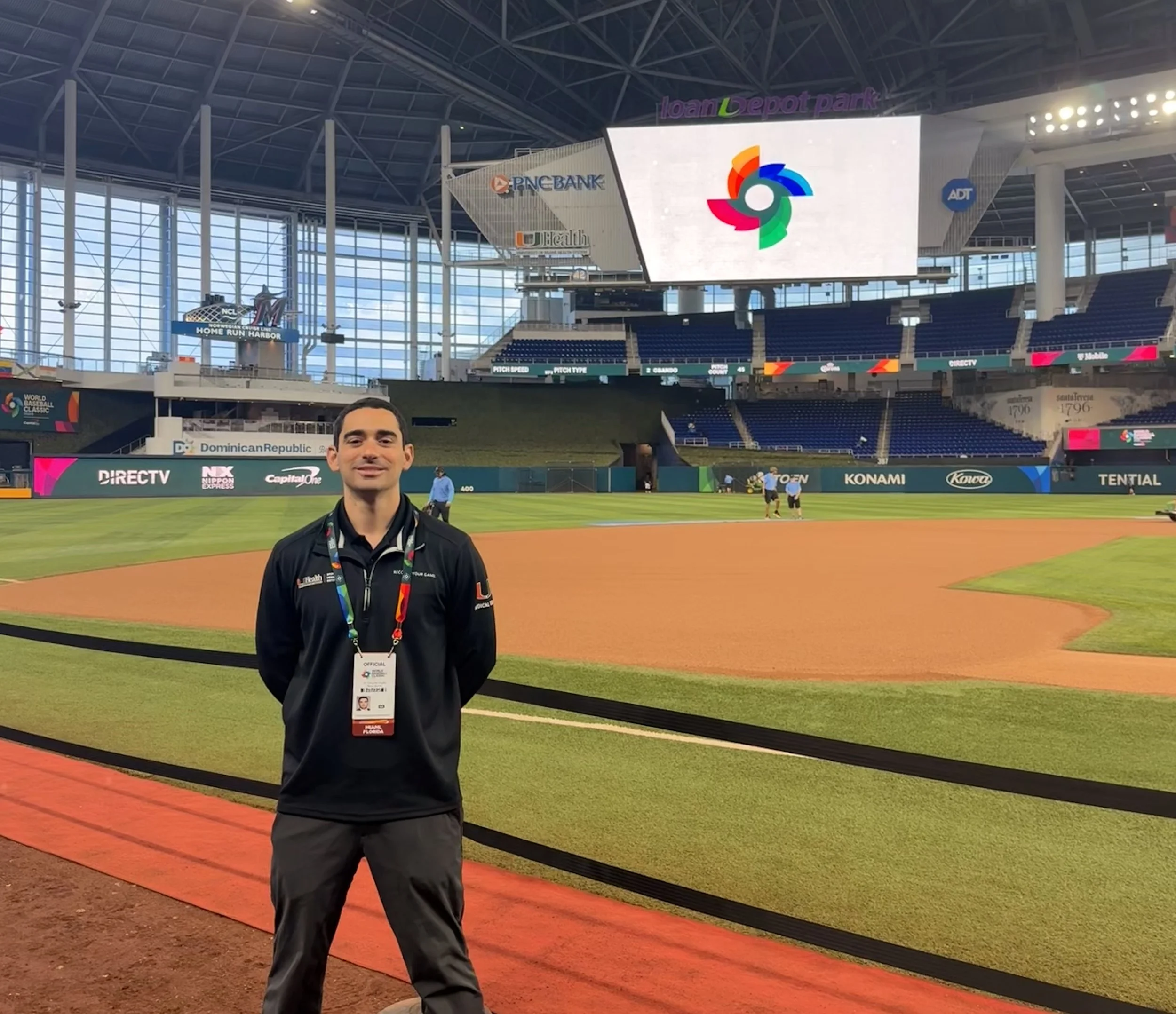A man smiling on a baseball field with large digital screens in the background showing logos of different sponsors and organizations at a stadium.