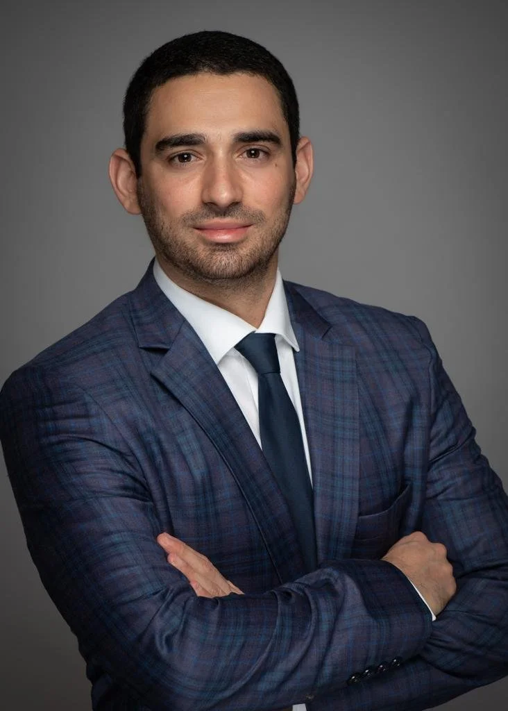 A man in a dark blue plaid suit with a white shirt and dark tie, posing with arms crossed against a gray background.
