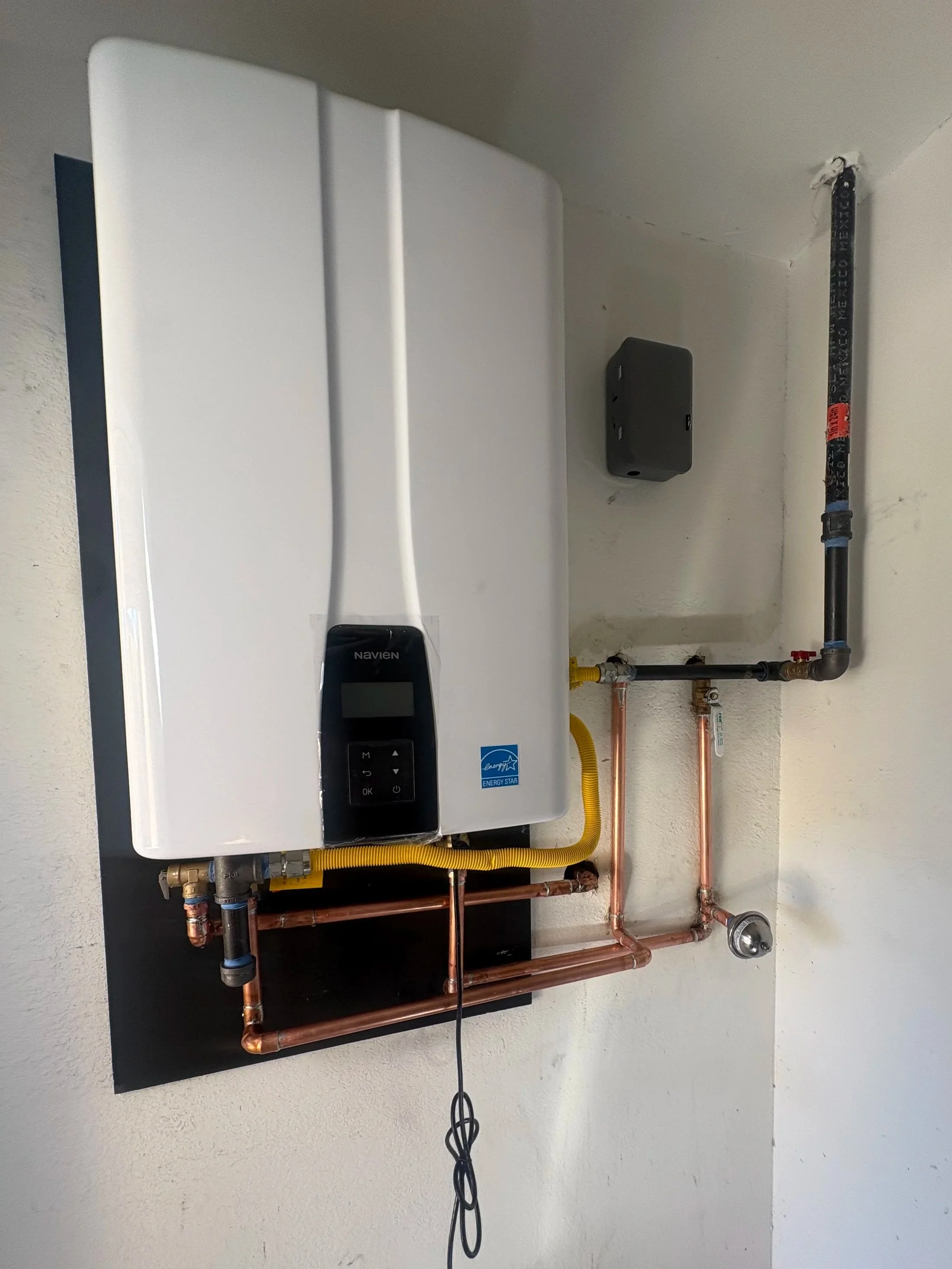 A white modern tankless water heater with digital controls is mounted on a white wall, connected to copper and black pipes, with a black remote control attached by a black cord hanging below.