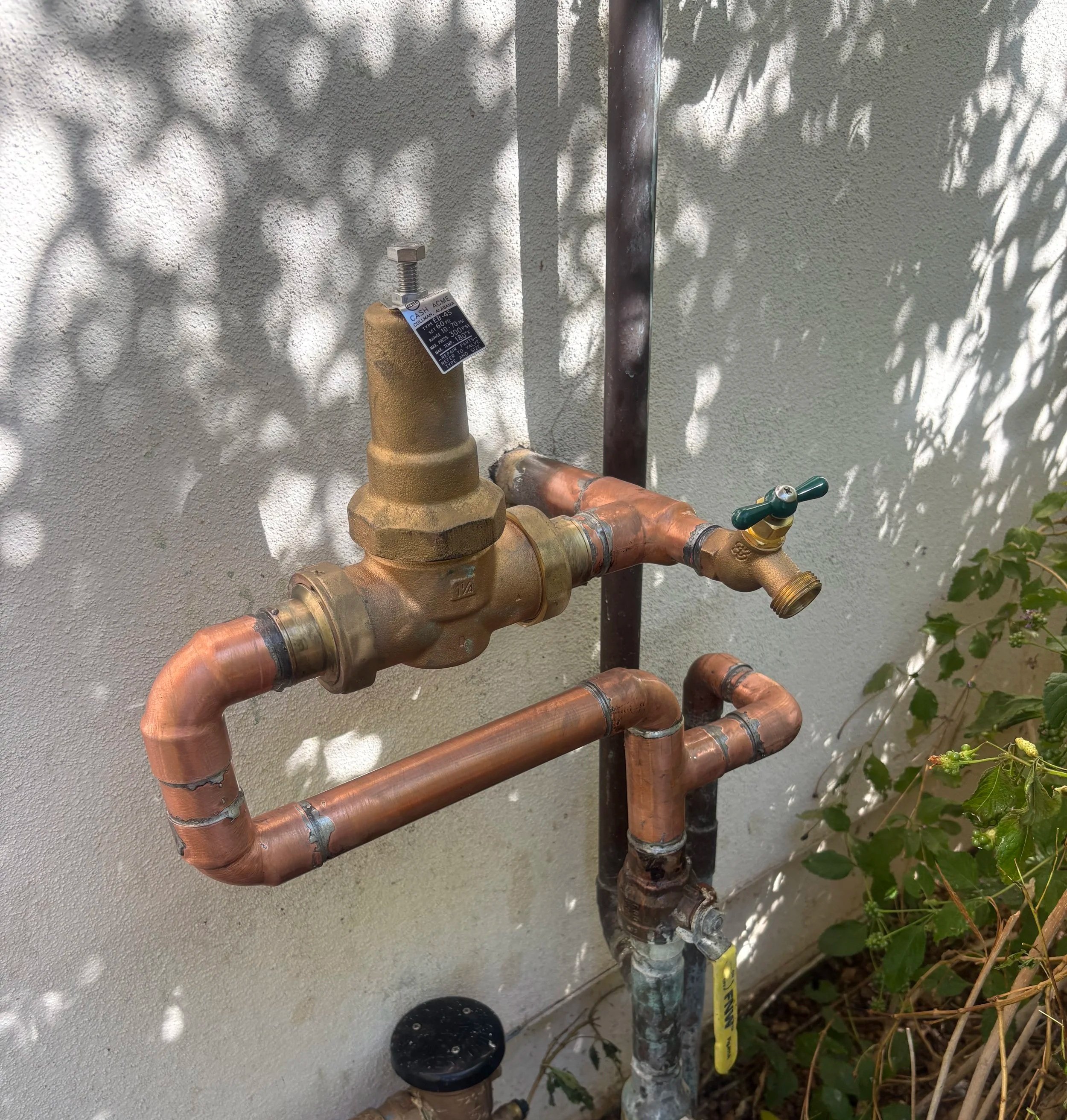 Close-up of copper and brass plumbing pipes and valves on a textured white wall, with a shadow of leaves cast on the wall.