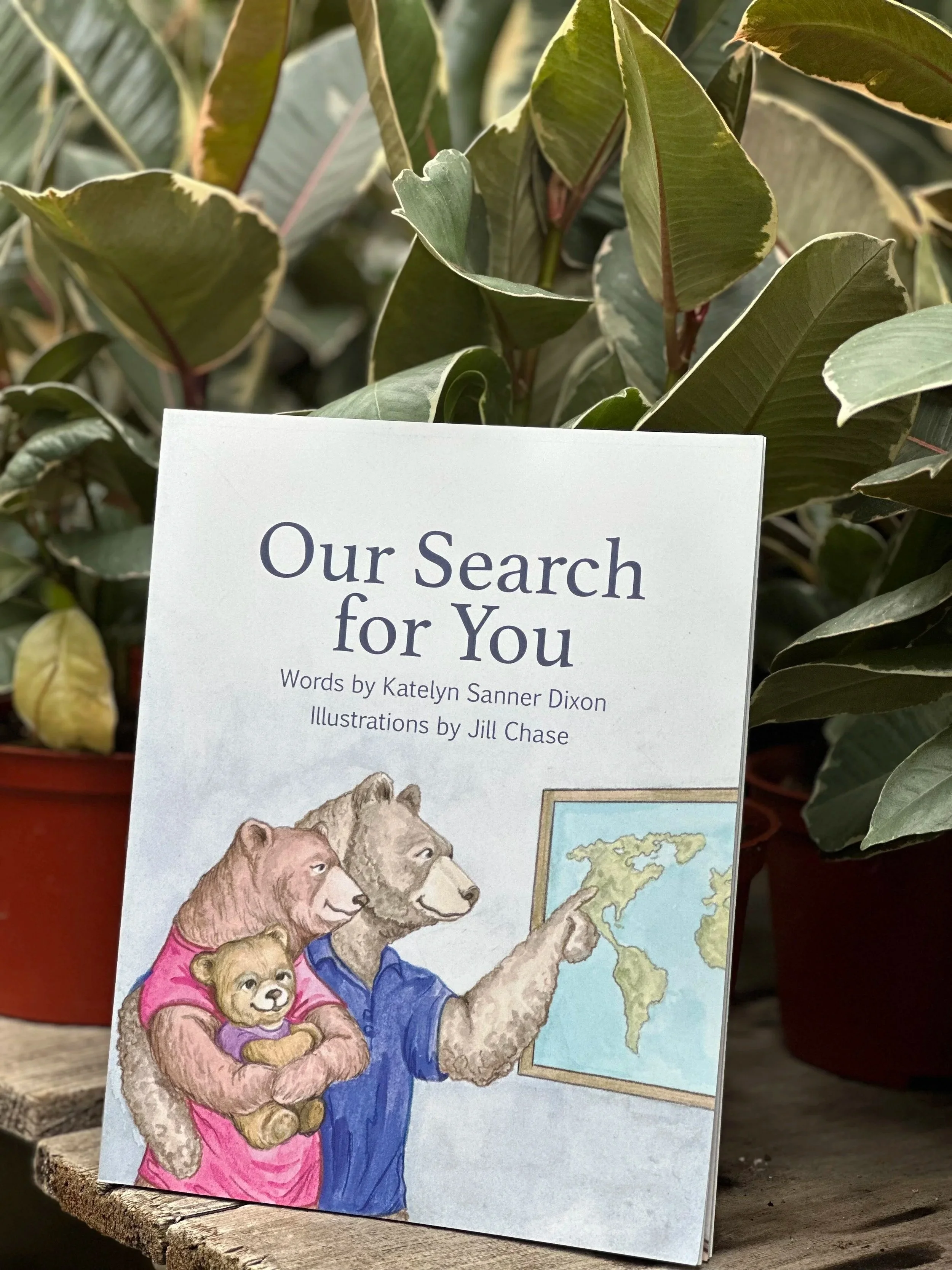 Children's book titled 'Our Search for You' with illustrations of animals at a map, placed in front of leafy plants.