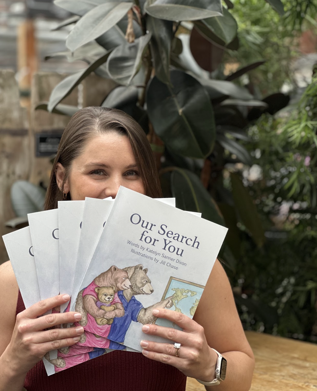 A woman holding multiple copies of a children's book titled 'Our Search for You' with an illustration of animals in front of a large leafy plant.