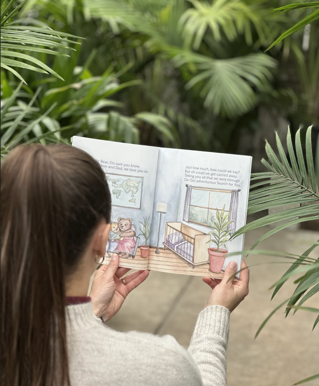 Person reading a children's picture book with a bear illustration, indoors near green tropical plants.