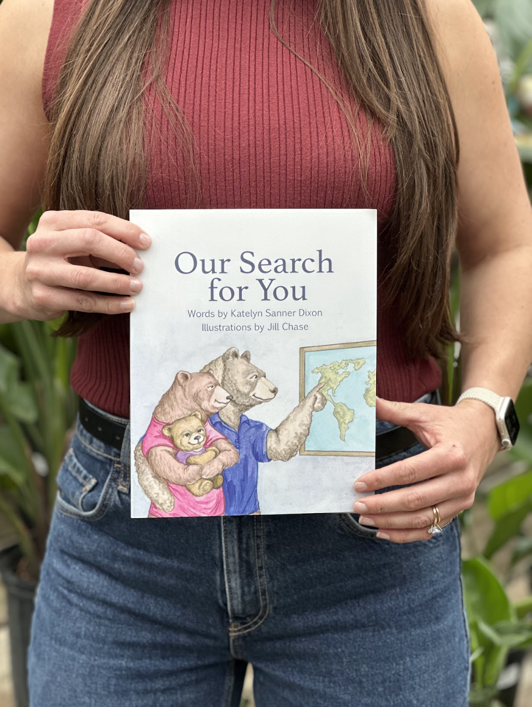 A person in a sleeveless red top and jeans holding a children's book titled 'Our Search for You' with illustrations of anthropomorphic bears and a world map on the cover.