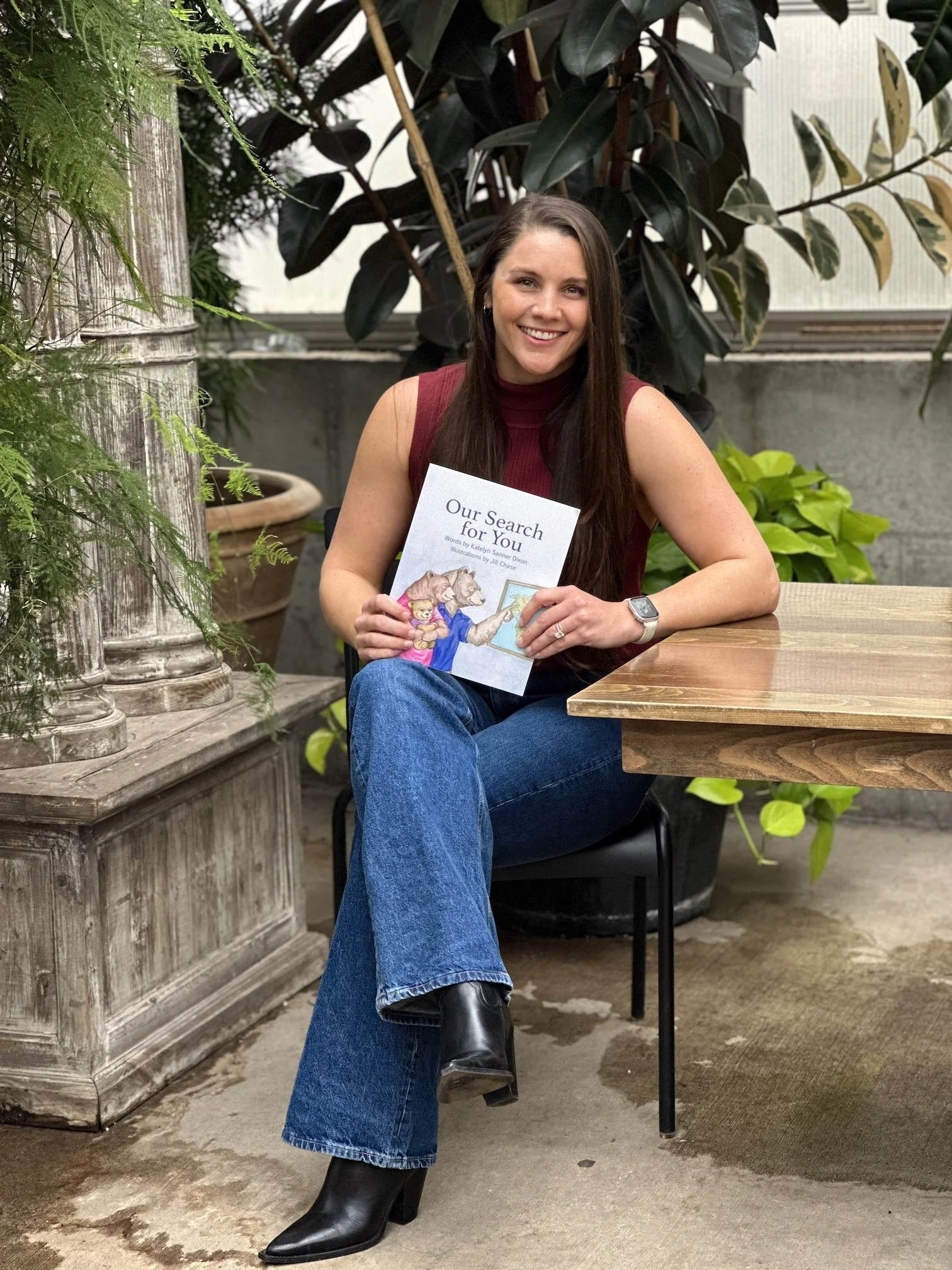 A woman with long dark hair, wearing a sleeveless burgundy top, blue jeans, and black boots, sitting at a wooden table in a greenhouse or botanical setting, smiling and holding a children's book titled 'Our Search for You'.
