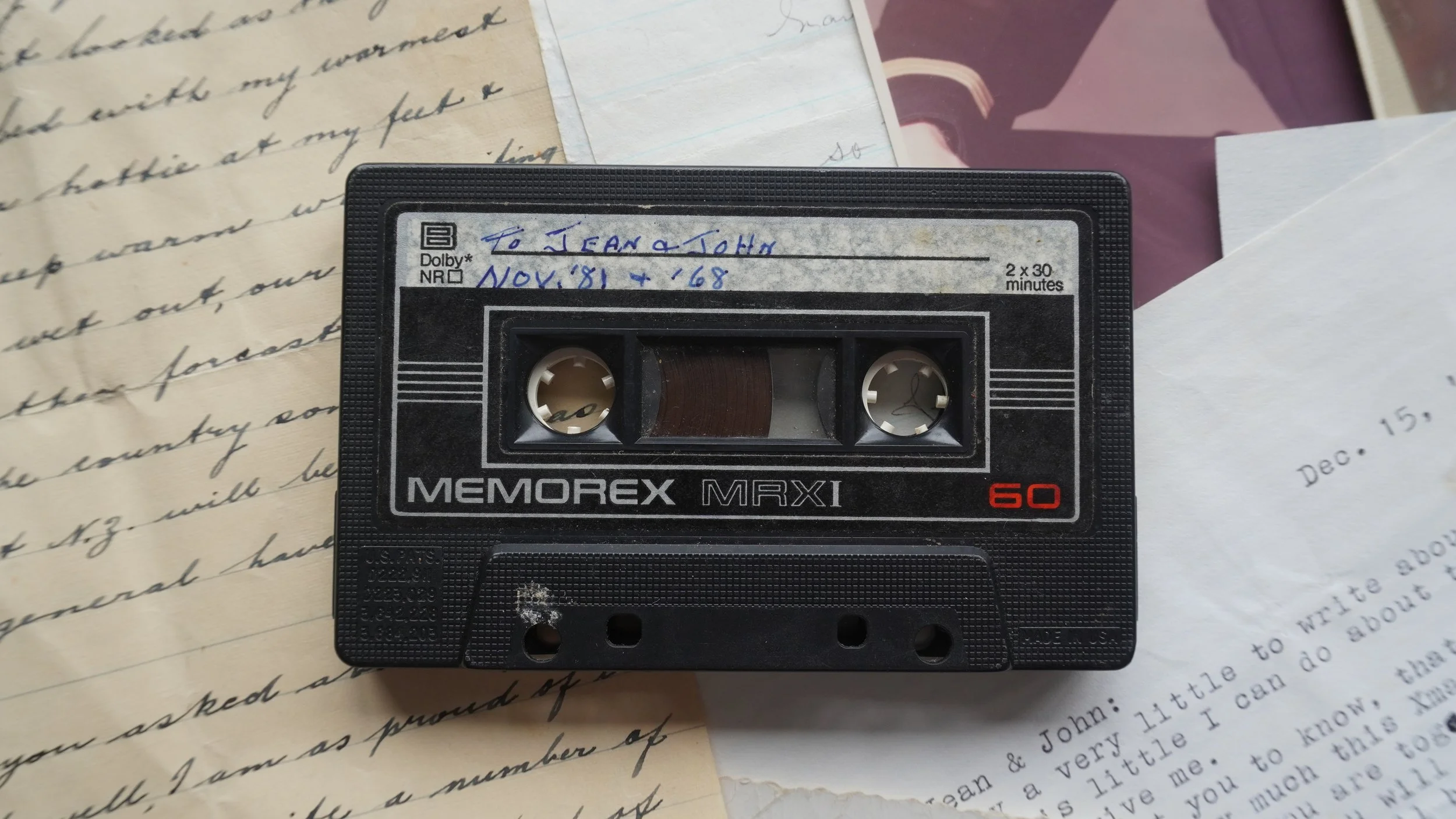 A black Memorex MRX1 audio cassette tape with a handwritten label on top, placed on scattered pieces of handwritten and typed paper documents.