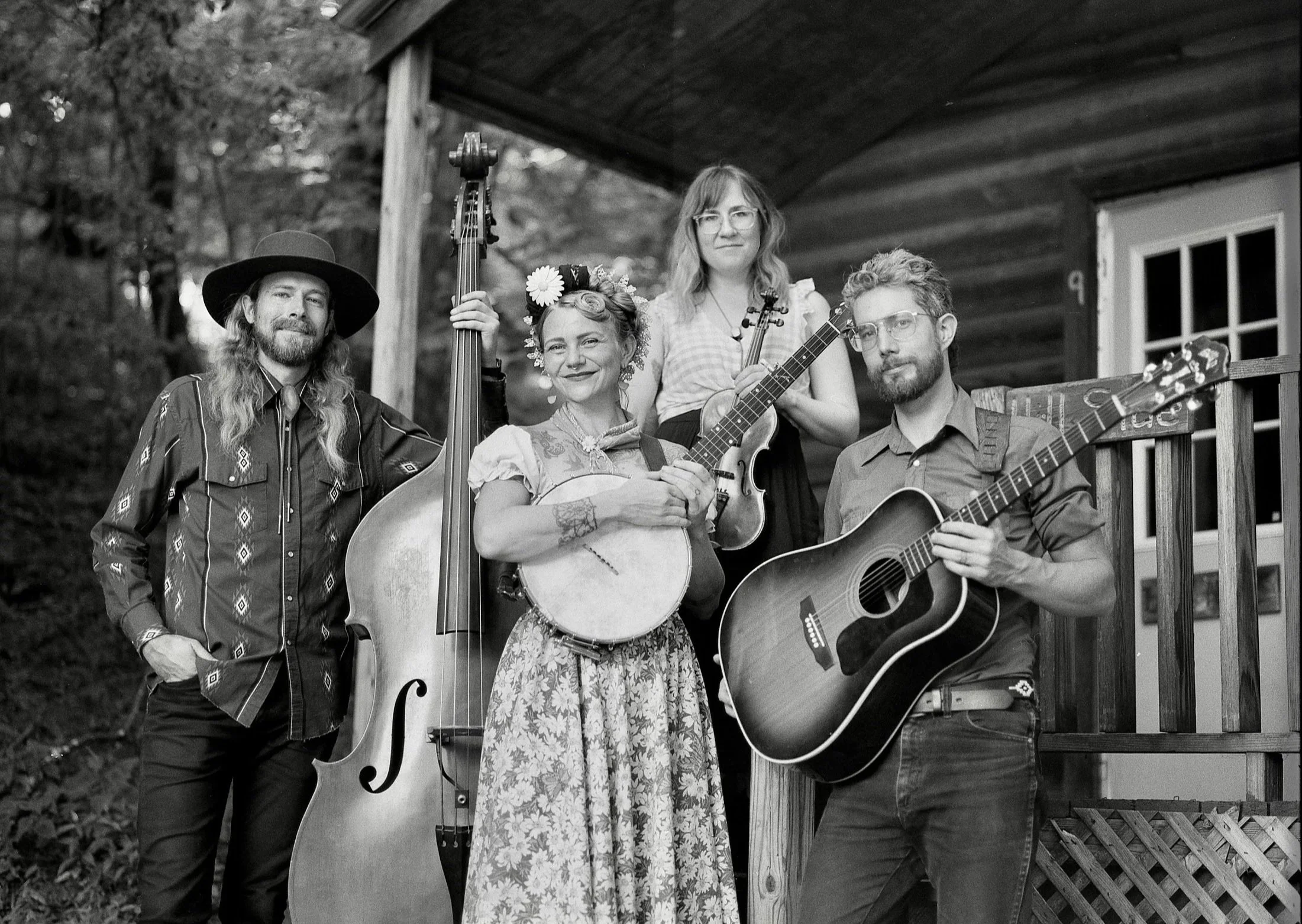 Five musicians holding string instruments on a porch, with a wooded background.