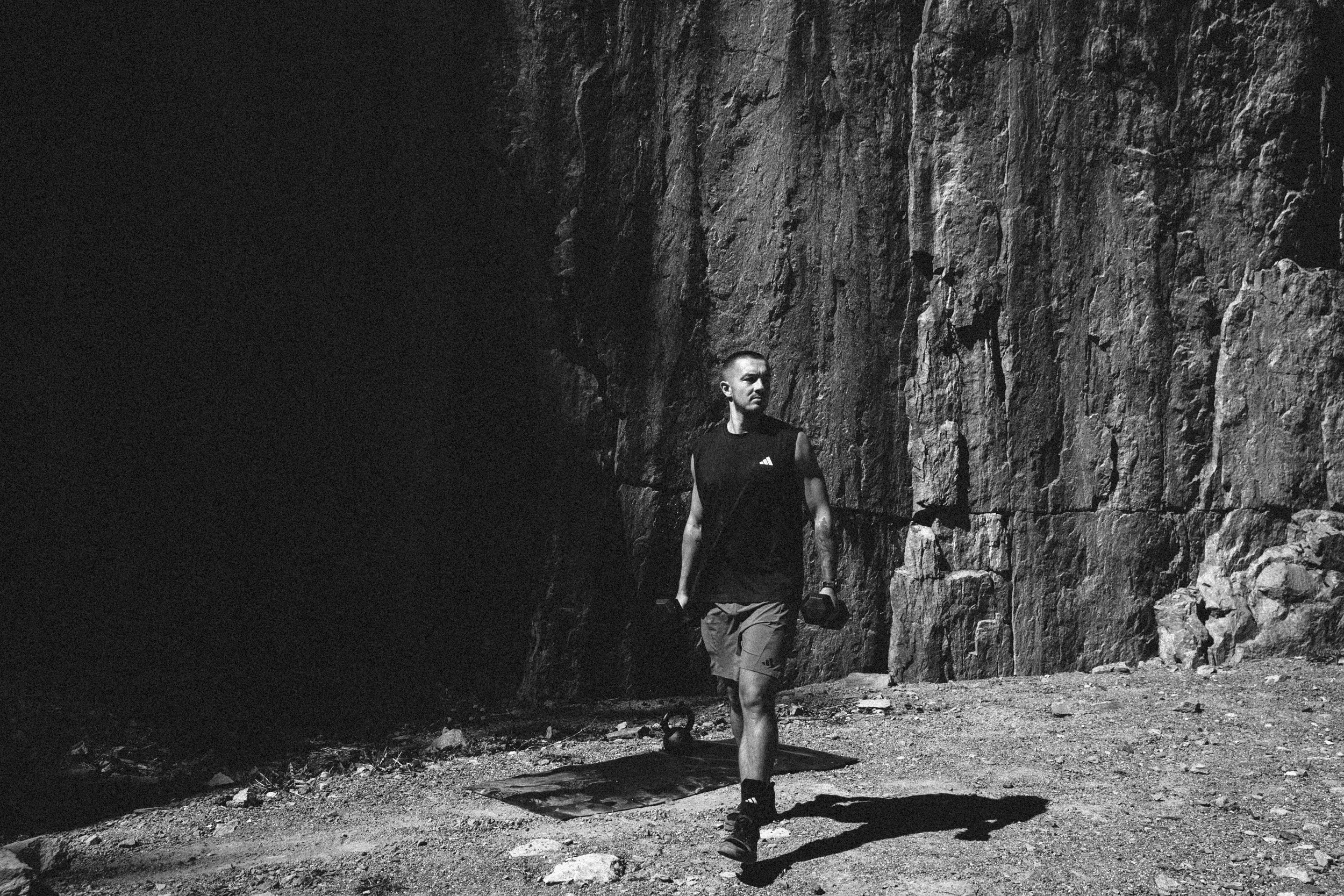 A man in a sleeveless shirt and shorts holding a kettlebell in each hand walking outdoors in front of a rock cliff.