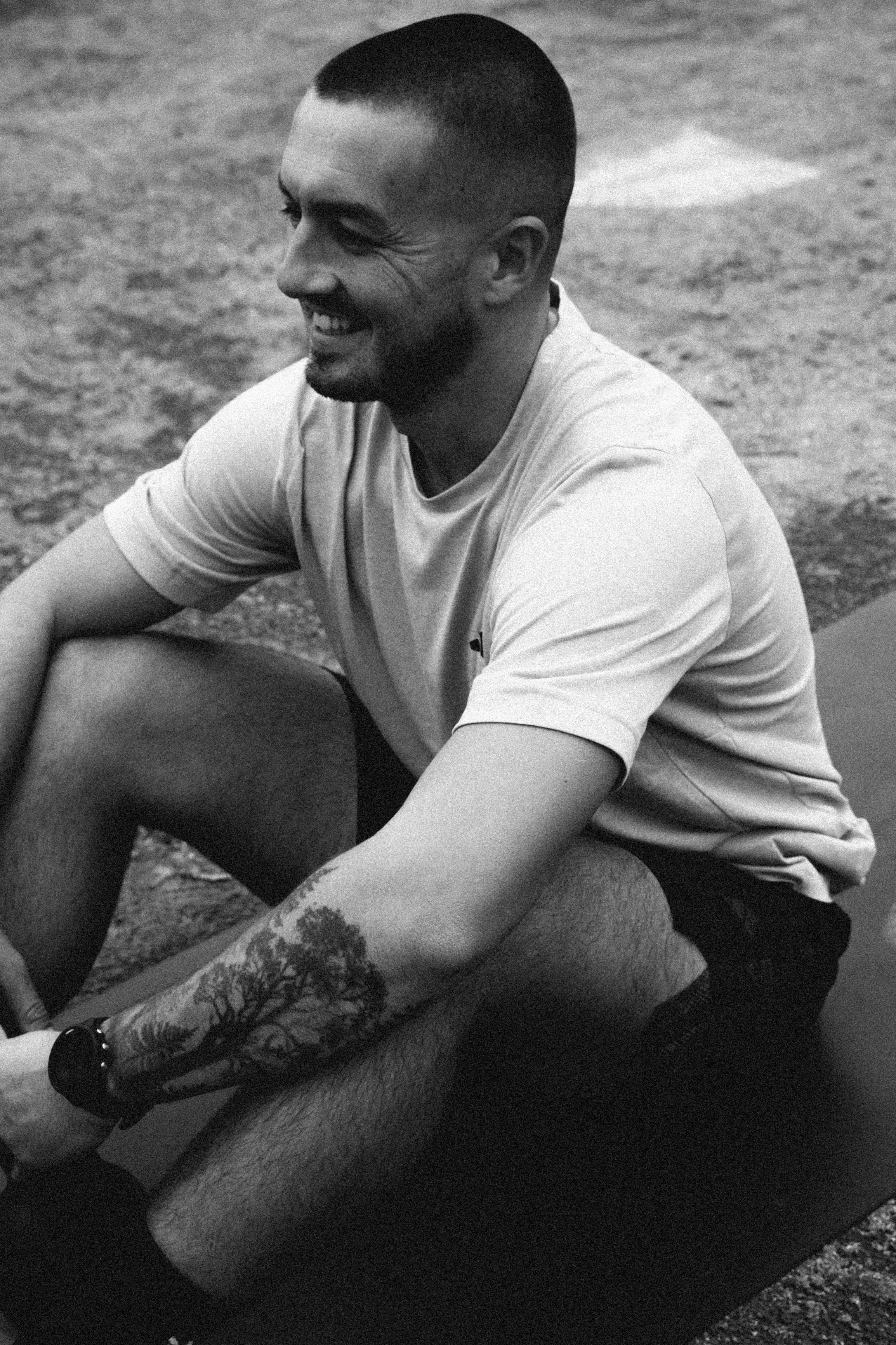 A smiling man with a short haircut and a beard, wearing a light-colored T-shirt, sitting on the ground with legs crossed. His arm and leg are tattooed with patterns. The background appears to be an outdoor surface.