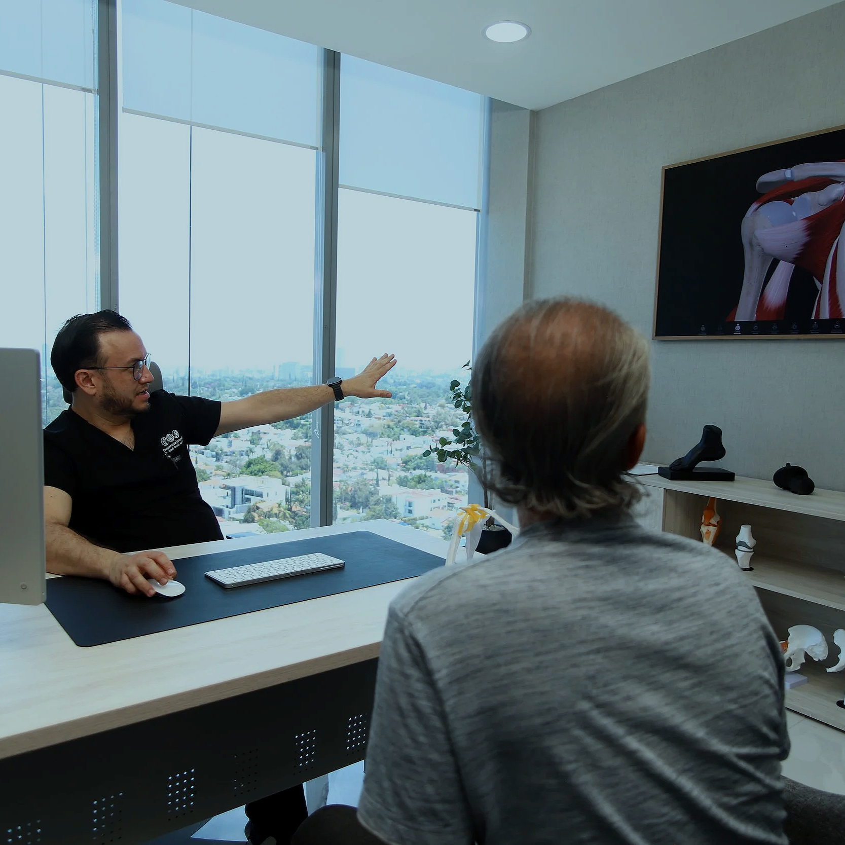 Reunión en oficina moderna con tres personas, una de ellas explicando con gestos frente a una pantalla con músculos del cuerpo humano, grandes ventanas con vista urbana de fondo.