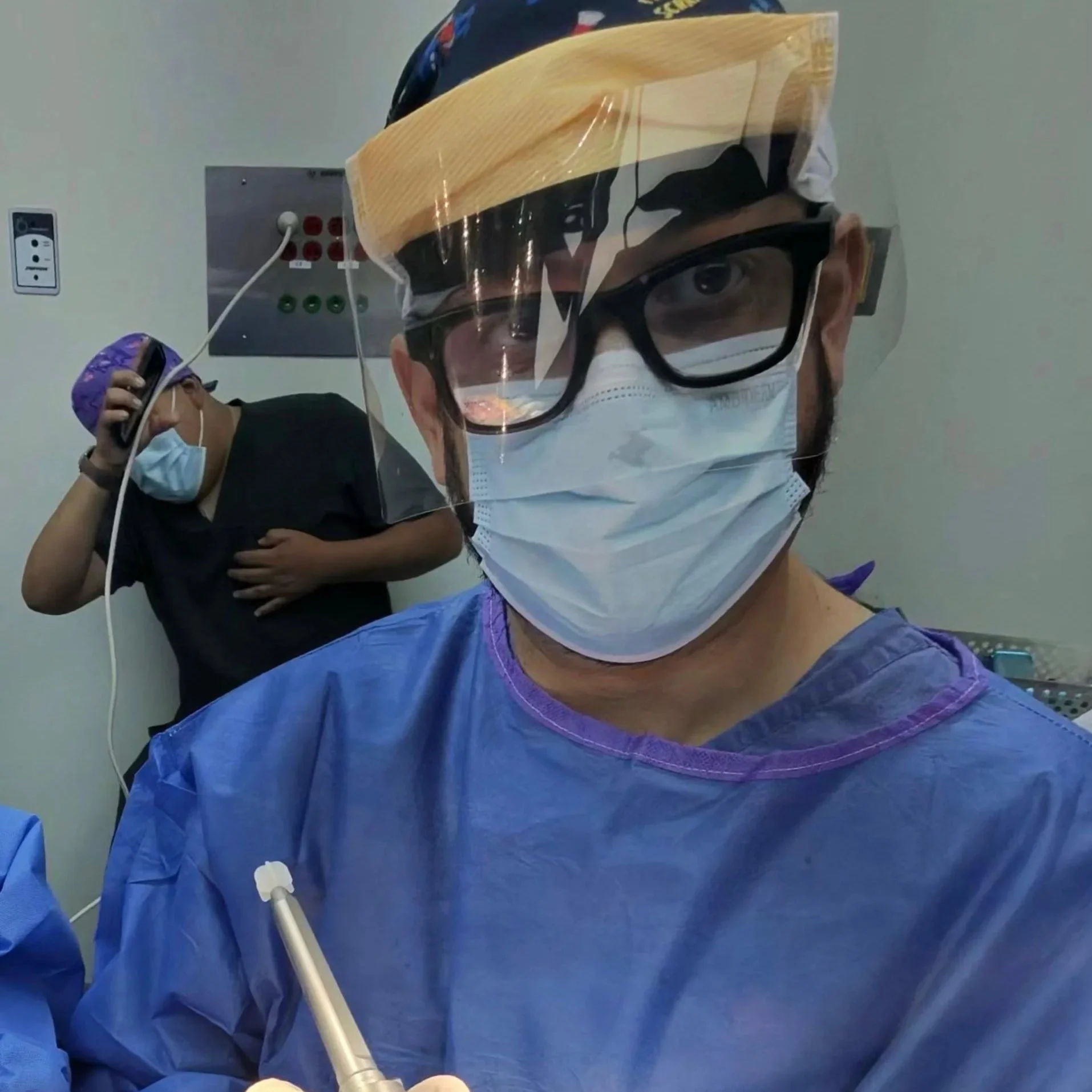 El Dr. Israel Romo ortopedista con protección completa en un hospital, en primer plano, y otro personal médico de fondo en una cirugia de revisión de rodilla