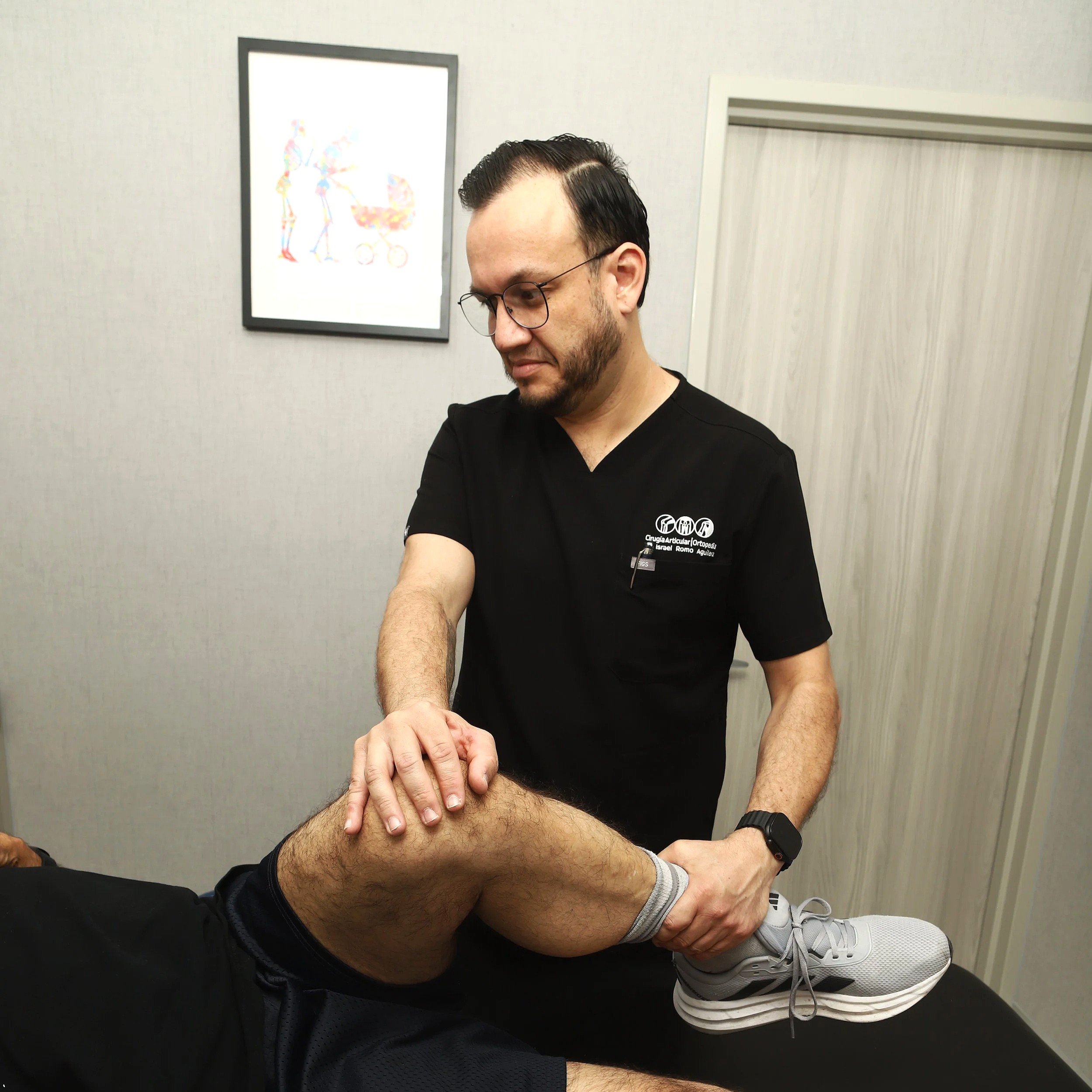 Un hombre con barba y gafas sentado en una sala, con un fondo de pared clara y un cuadro colorido, sosteniendo la pierna de otra persona que lleva pantalón gris y tenis blancos.
