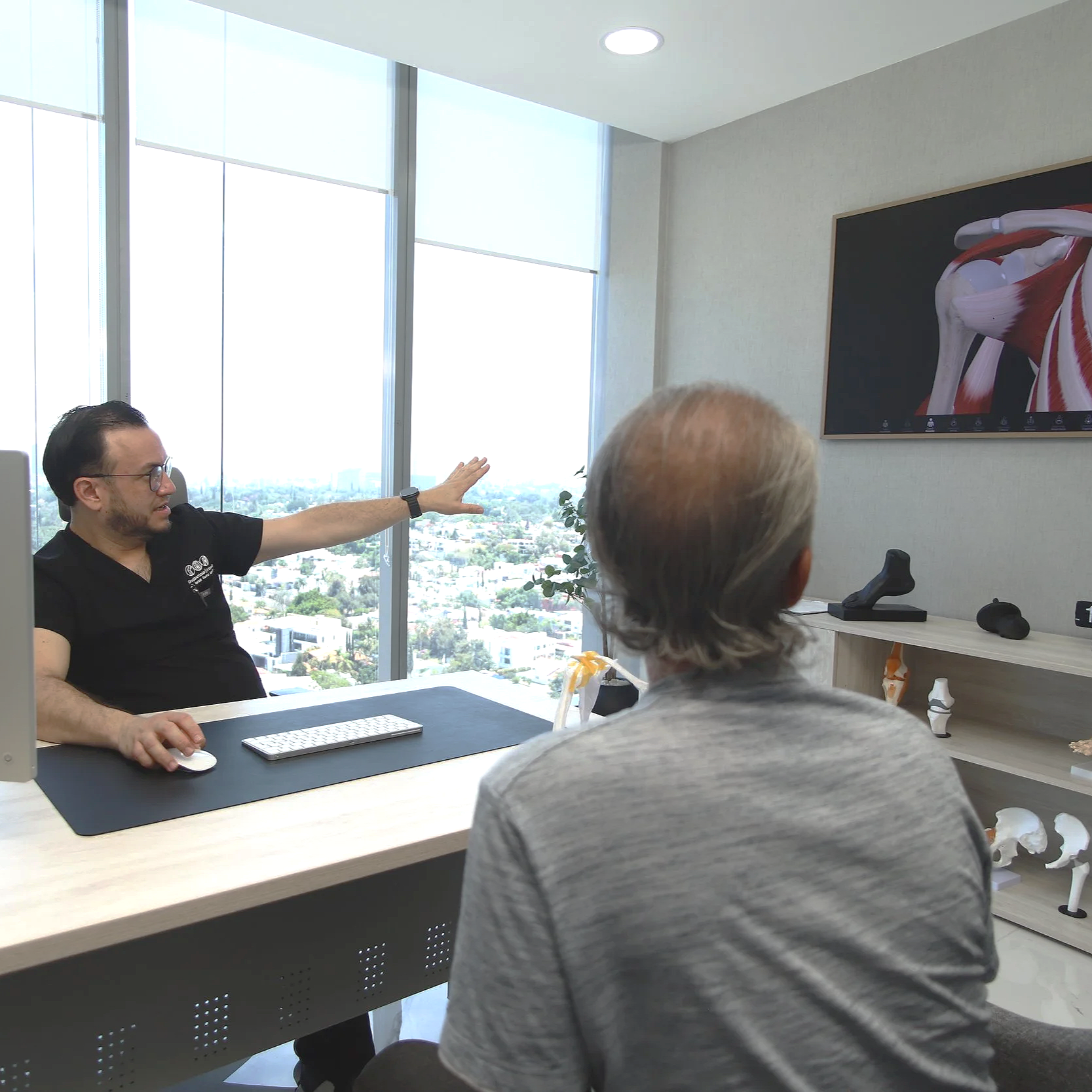 Una persona en una oficina de aspecto moderno, presenta a una audiencia a través de videoconferencia, con un gran monitor que muestra la parte superior de un músculo del cuerpo humano, en un entorno con ventanas grandes y vista panorámica de la ciudad.
