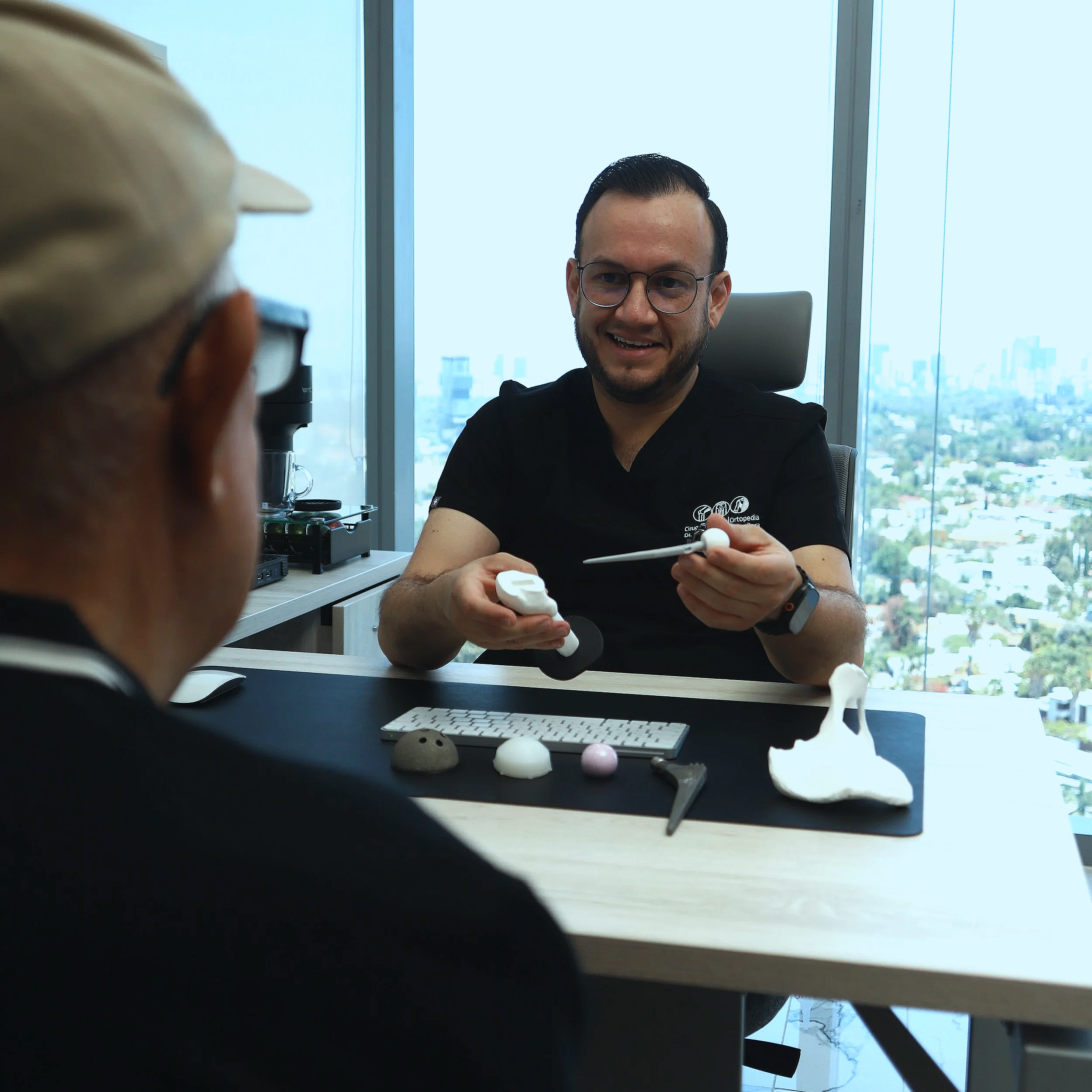 Hombre con gafas sosteniendo un modelo de hueso, sentado en una oficina con grandes ventanas y vista a la ciudad.