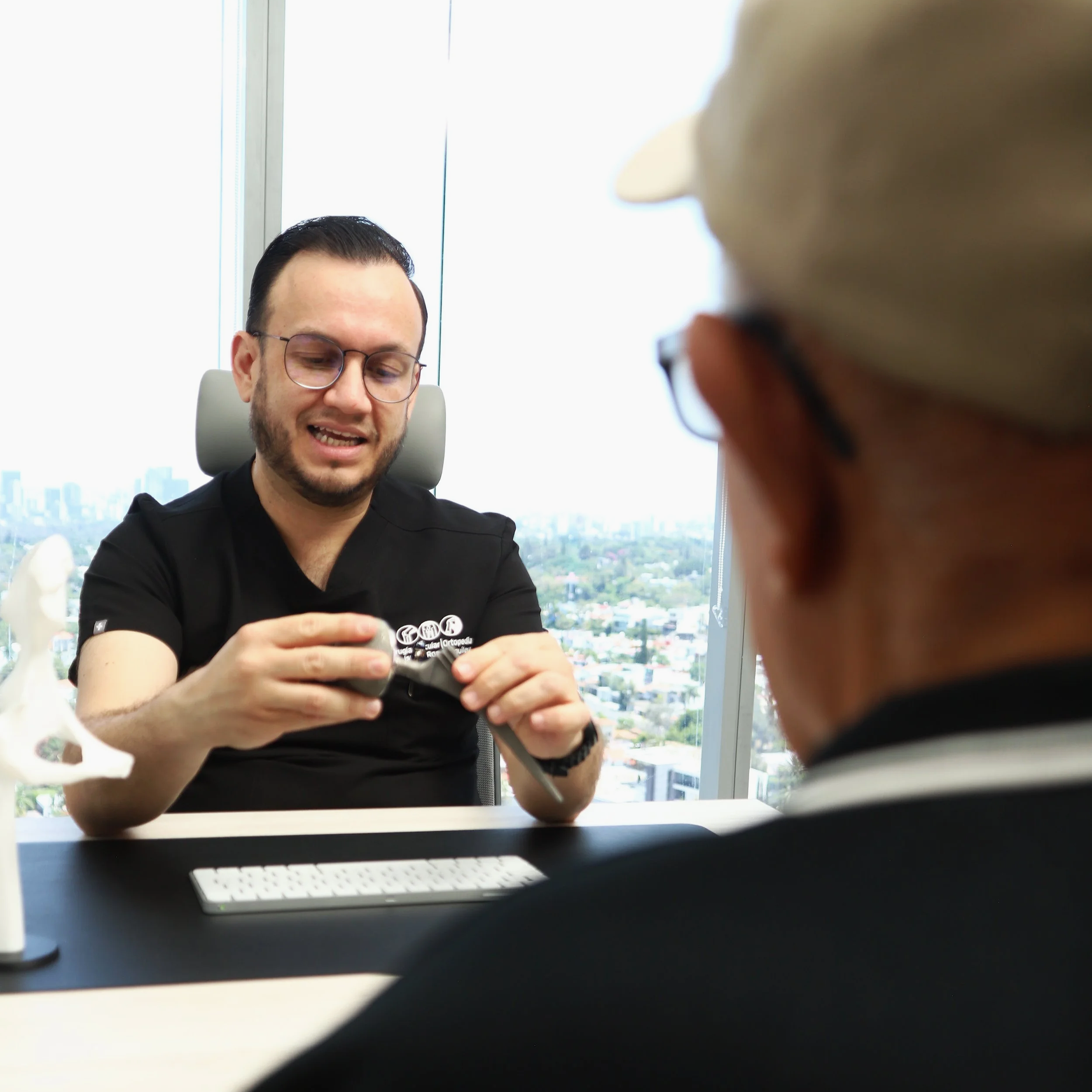 Hombre con gafas sentado en una oficina con vista de ciudad, sosteniendo un objeto en las manos, vista parcial de otra persona en primer plano en el mismo espacio.