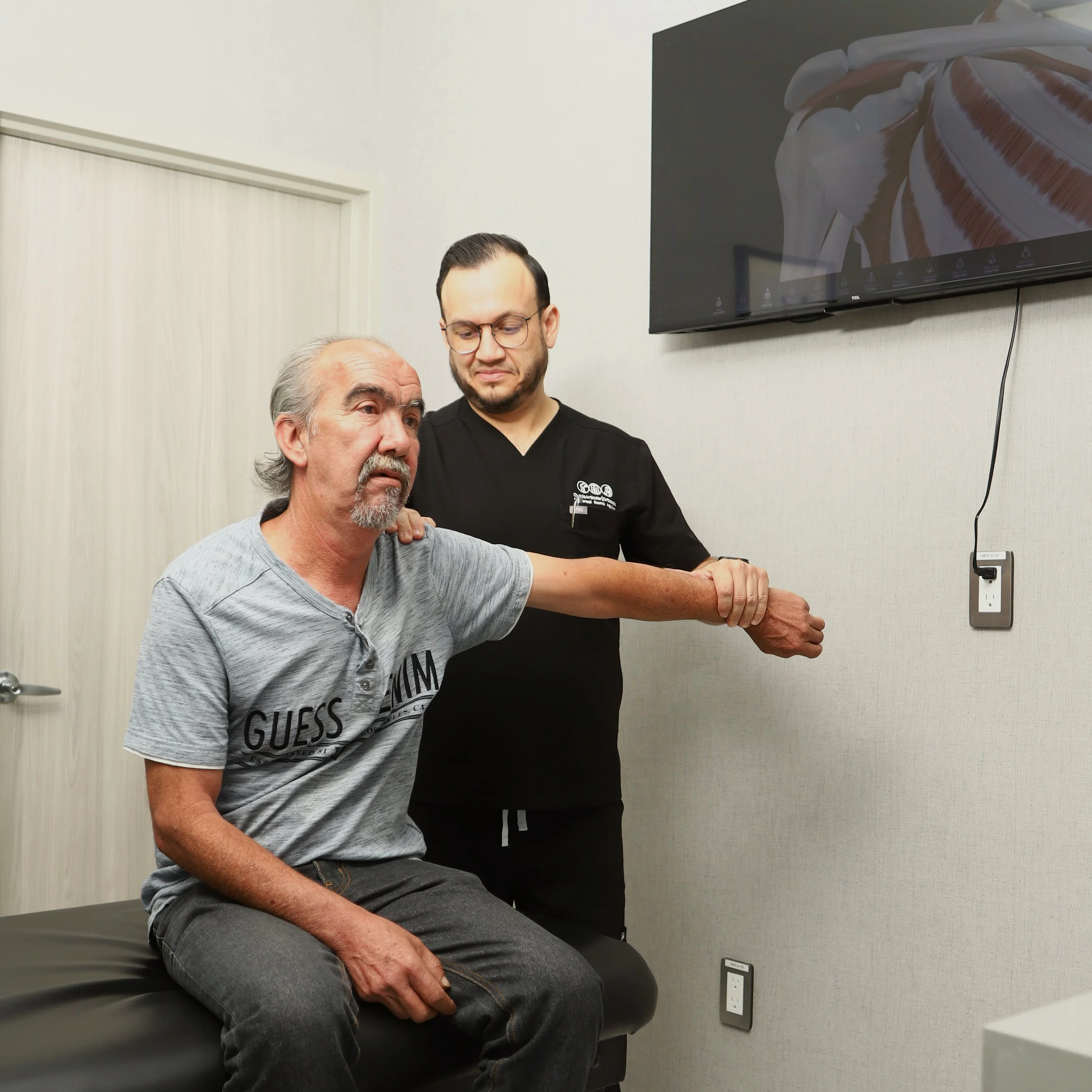 Un hombre mayor sentado en una camilla de exploración médica con un médico joven que lo examina, en una sala de consulta con pared de color claro y una televisión montada en la pared.