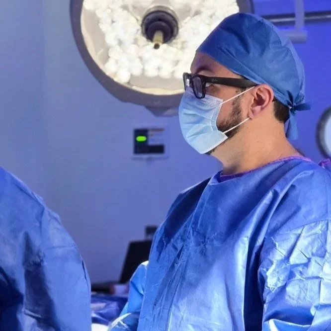 Dr. Israel Romo en cirugía ortopédica, médico en quirófano equipado con bata, gorro, mascarilla y gafas, realizando una cirugía.