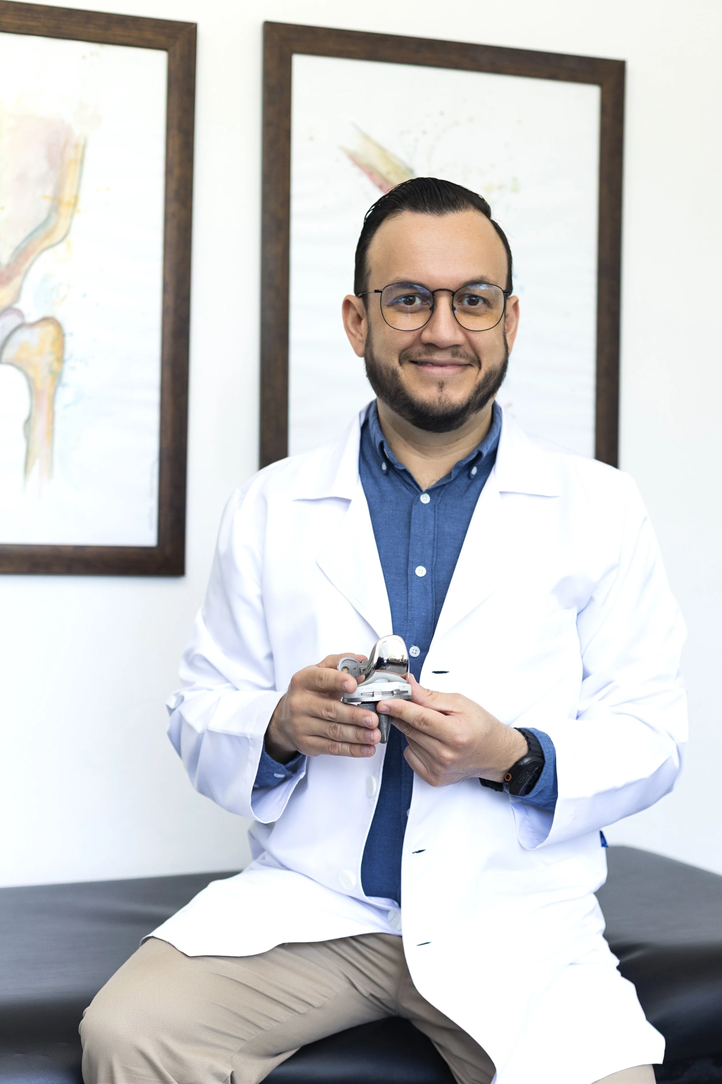 Un hombre con barba, gafas, camiseta azul y bata blanca, sentado en una camilla en un consultorio, sonriendo y sosteniendo un estetoscopio.