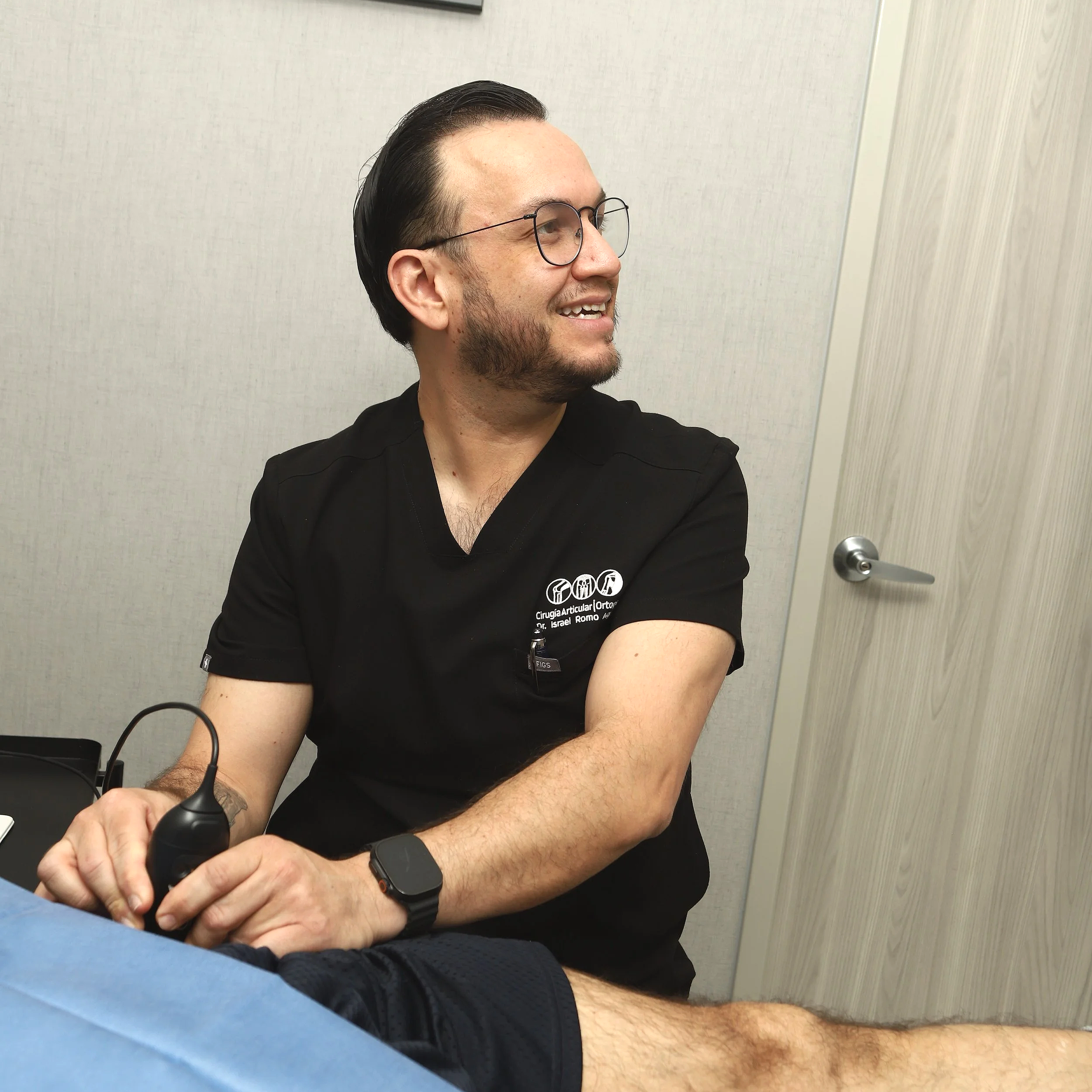 Hombre con gafas y barba sonriendo, sentado en una sala con paredes claras, usando una camiseta negra con texto y un reloj inteligente en la muñeca izquierda, sosteniendo un aparato en la mano derecha.