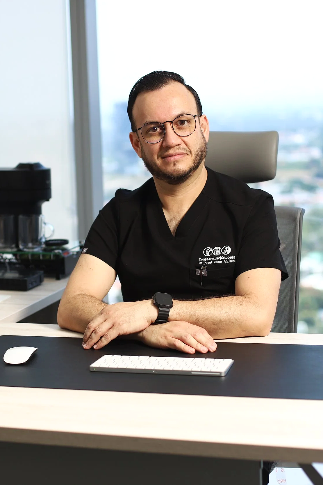 Hombre en oficina sentado en una mesa con teclado, ratón y una computadora portátil, usando gafas y camiseta negra con logotipo y texto de cirugía ortopédica, con vista de ciudad por la ventana detrás.