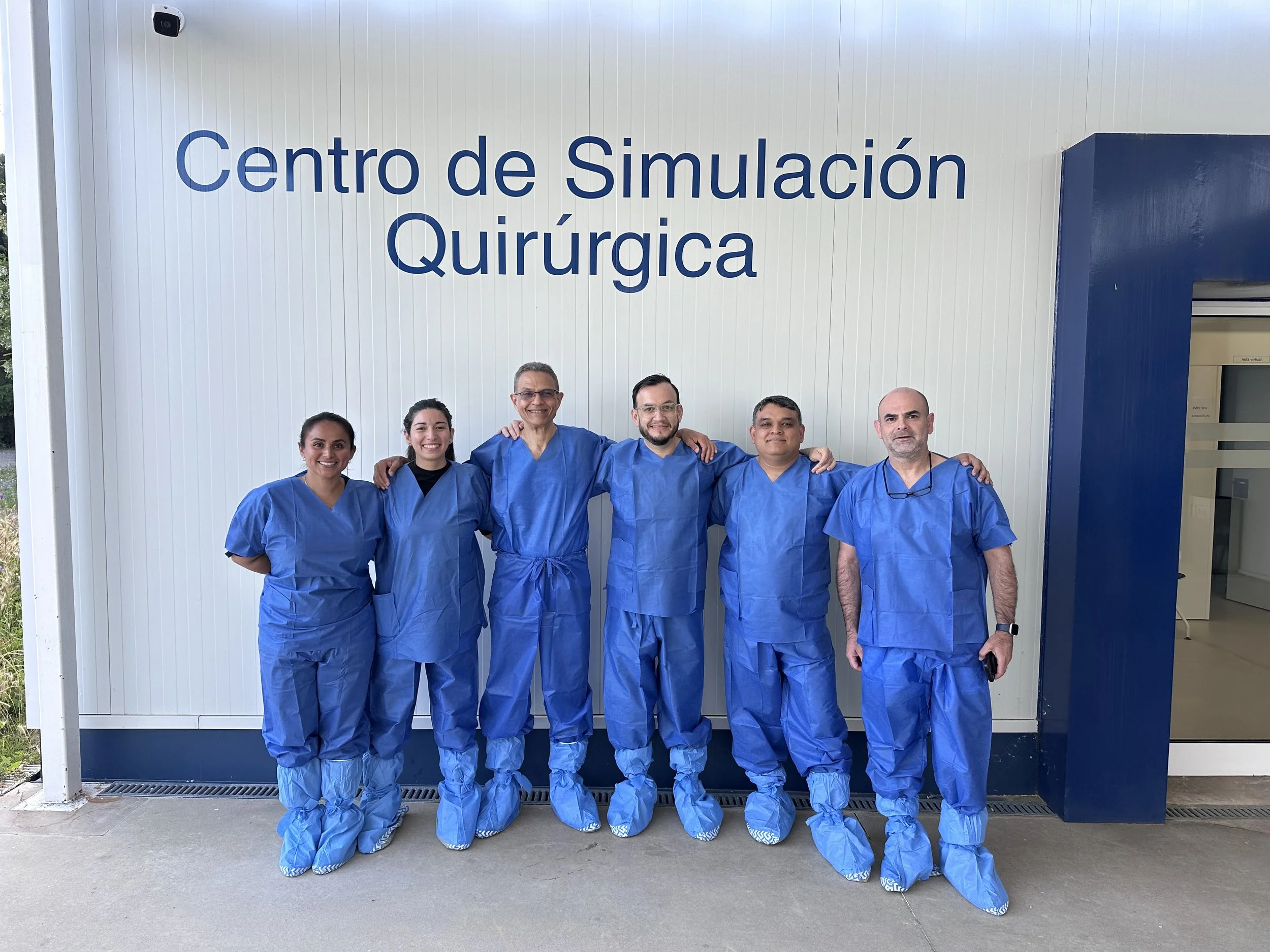 Grupo de seis profesionales en ropa de quirófano posando frente a una pared con un cartel que dice 'Centro de Simulación Quirúrgica'. Como parte de la formación médica del Dr. Israel Romo
