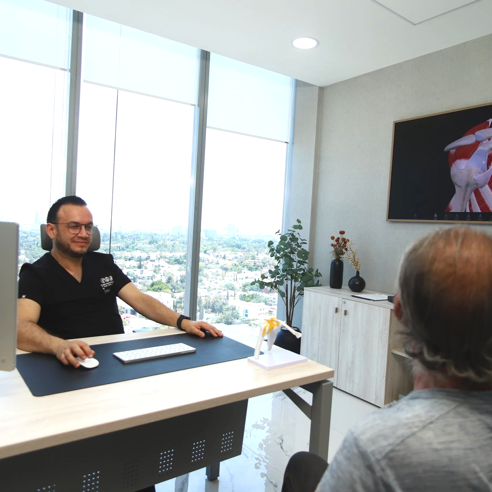 Hombre con gafas sentado en una oficina, participando en una reunión, con grandes ventanas y vista urbana en el fondo, y un cuadro de músculos en la pared.