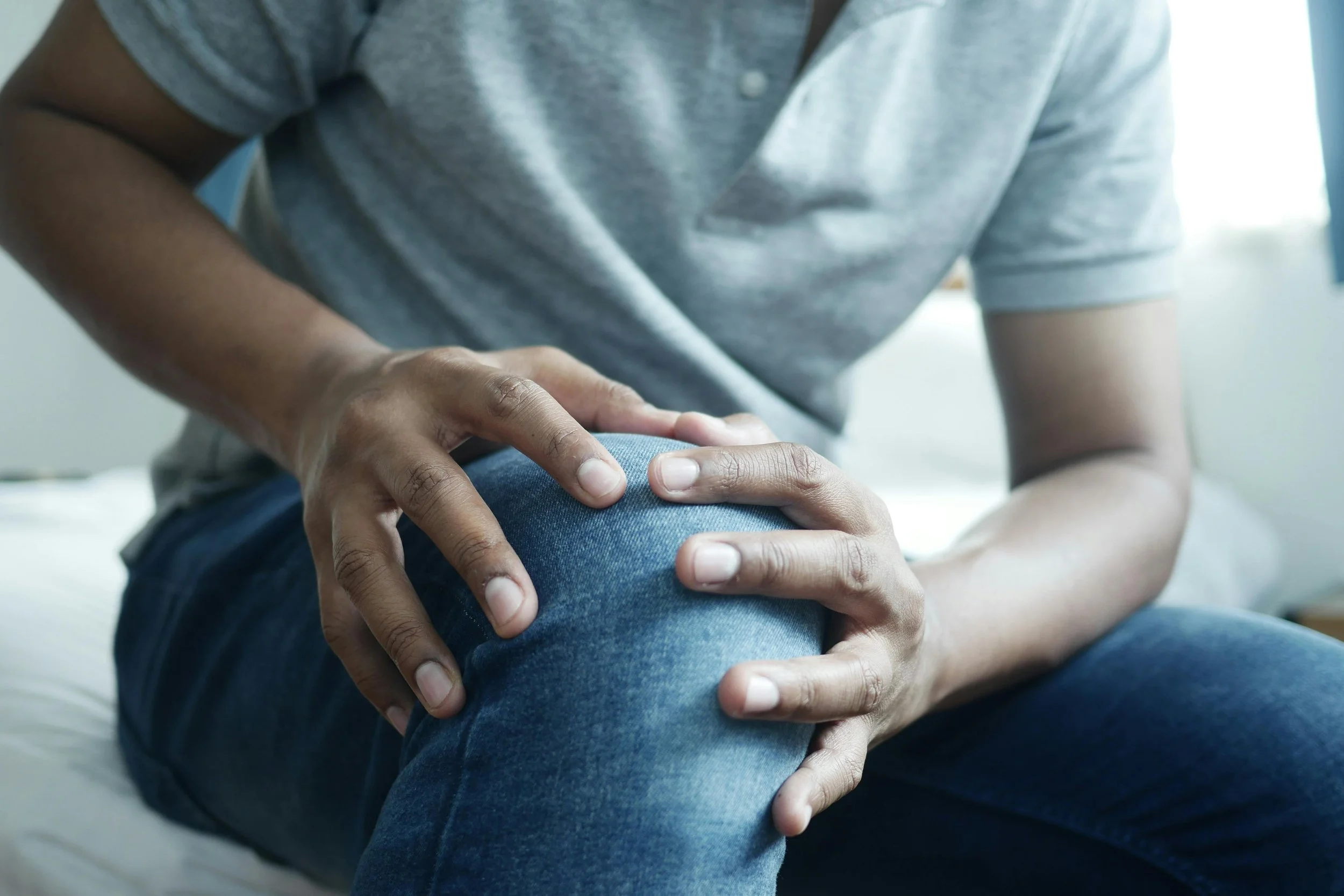 Persona sosteniendo su rodilla con ambas manos, en una postura de dolor o incomodidad, con una camisa gris y jeans azules.