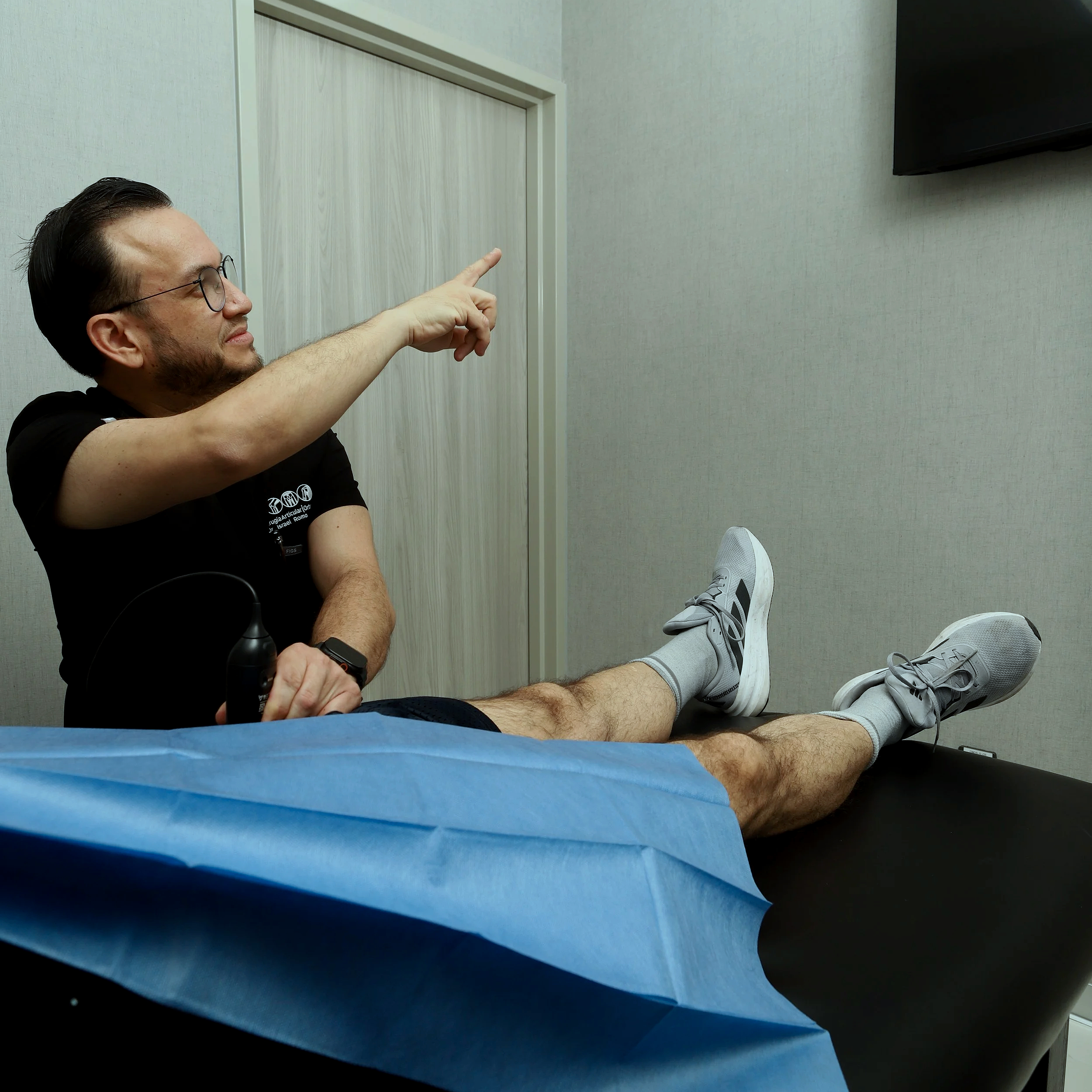 Un hombre con gafas y barba está recostado en una camilla en una sala sin ventanas, vestido con una camiseta negra con letras blancas, y le están colocando una aguja en el brazo. Tiene el cabello peinado hacia atrás y está mirando hacia la derecha mientras recibe una inyección.