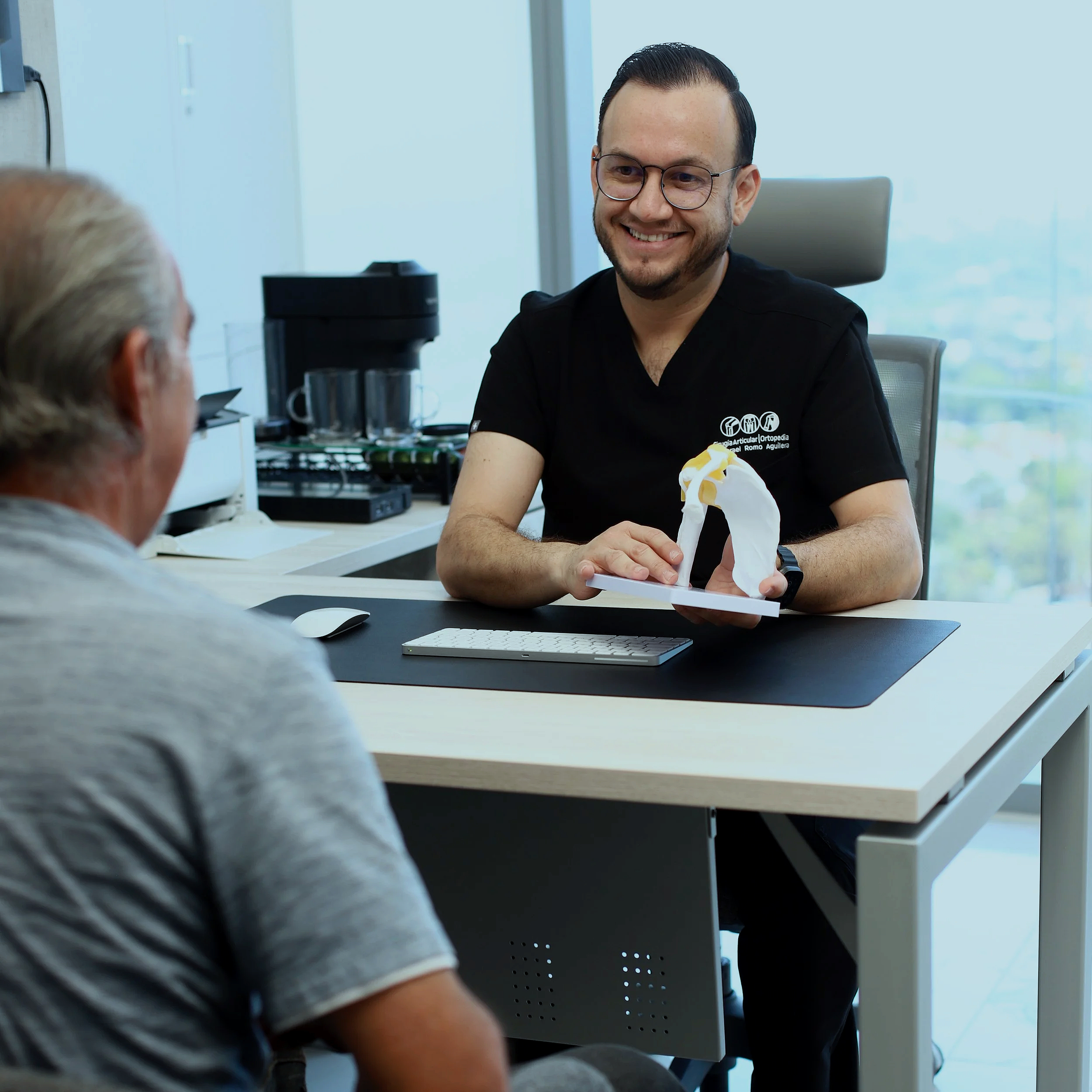 Un hombre con gafas y barba sonriendo en una oficina, sentado frente a una mujer con cabello gris que está parcialmente visible y una computadora en el escritorio.