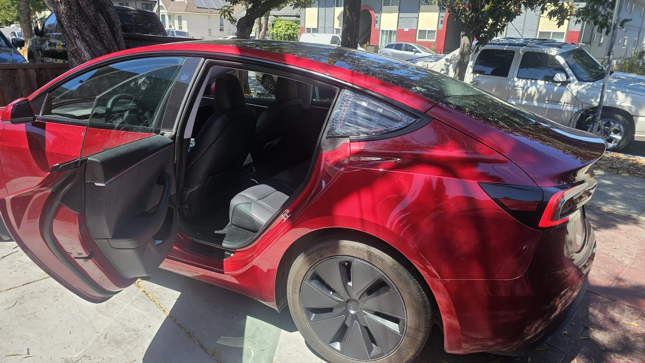 Red electric car parked on the street with open rear passenger door, showing black interior and rear seats, with residential buildings and trees in the background.