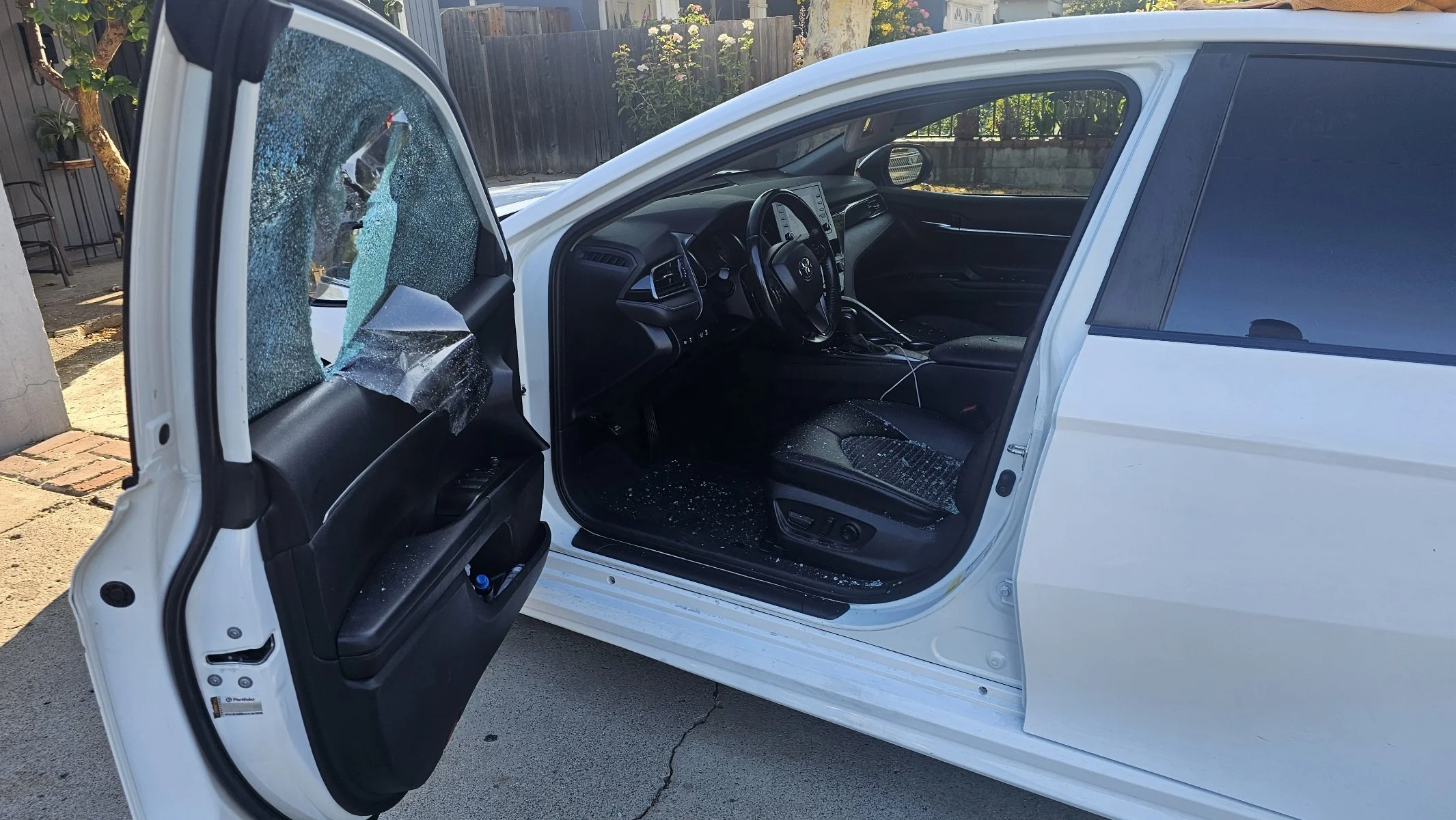 A white car with broken front and side windows, with glass shards on the driver's seat and floor, parked outdoors on a driveway.