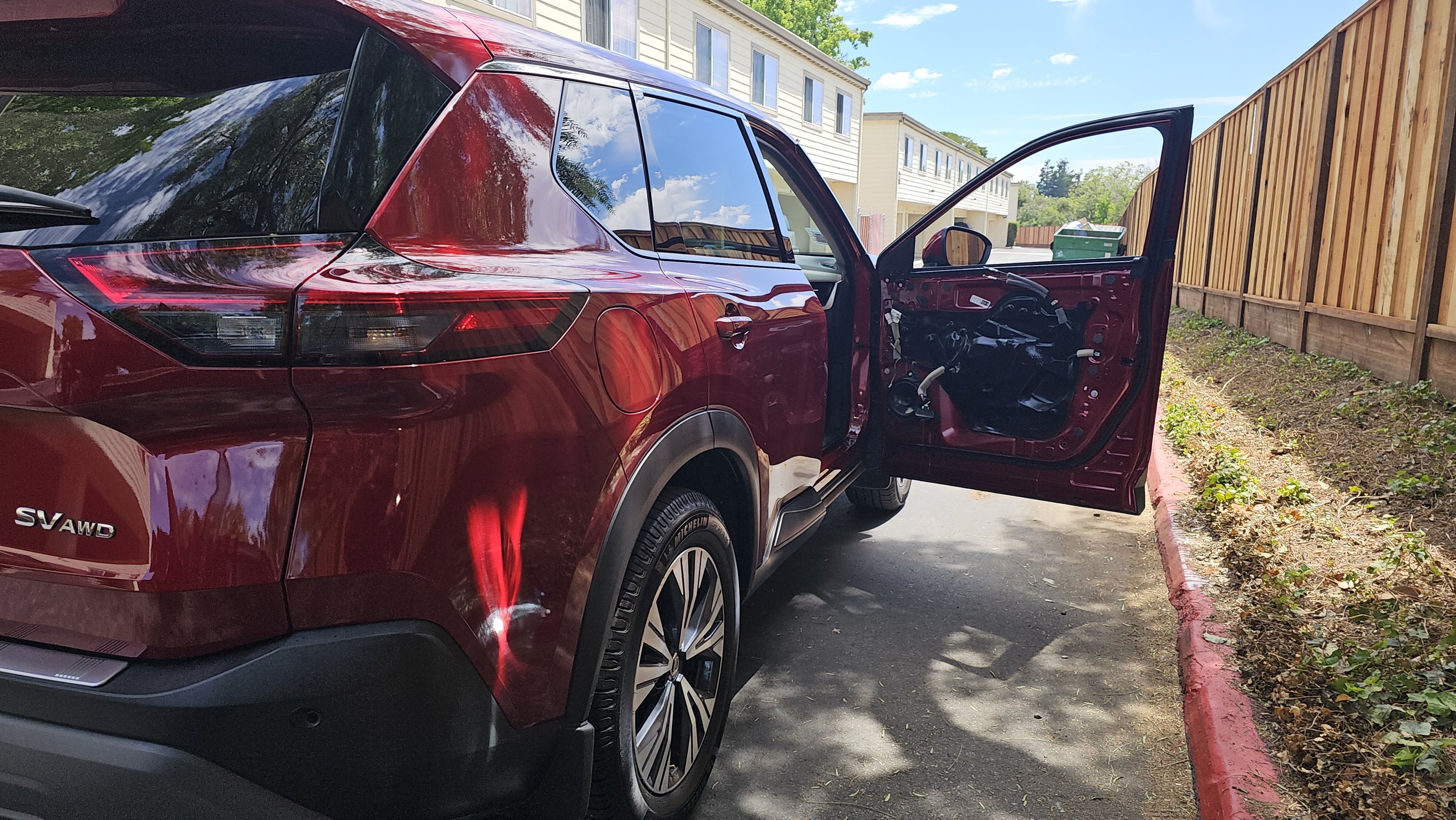 A red SUV with its front door open, parked on the side of a street near a wooden fence and residential buildings, with a trash bin visible in the background.