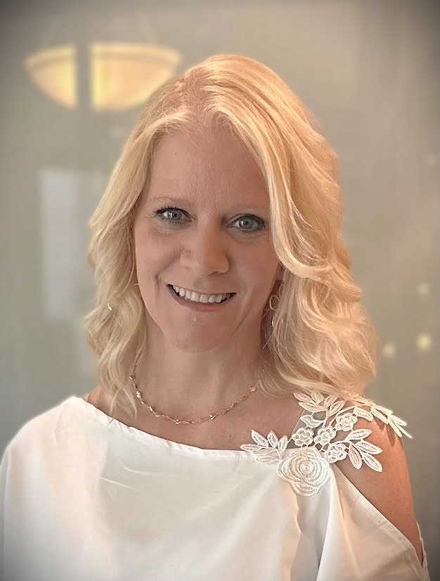 A woman with blonde hair styled in loose waves, wearing a white dress with floral embroidery on the shoulder, smiling at the camera.