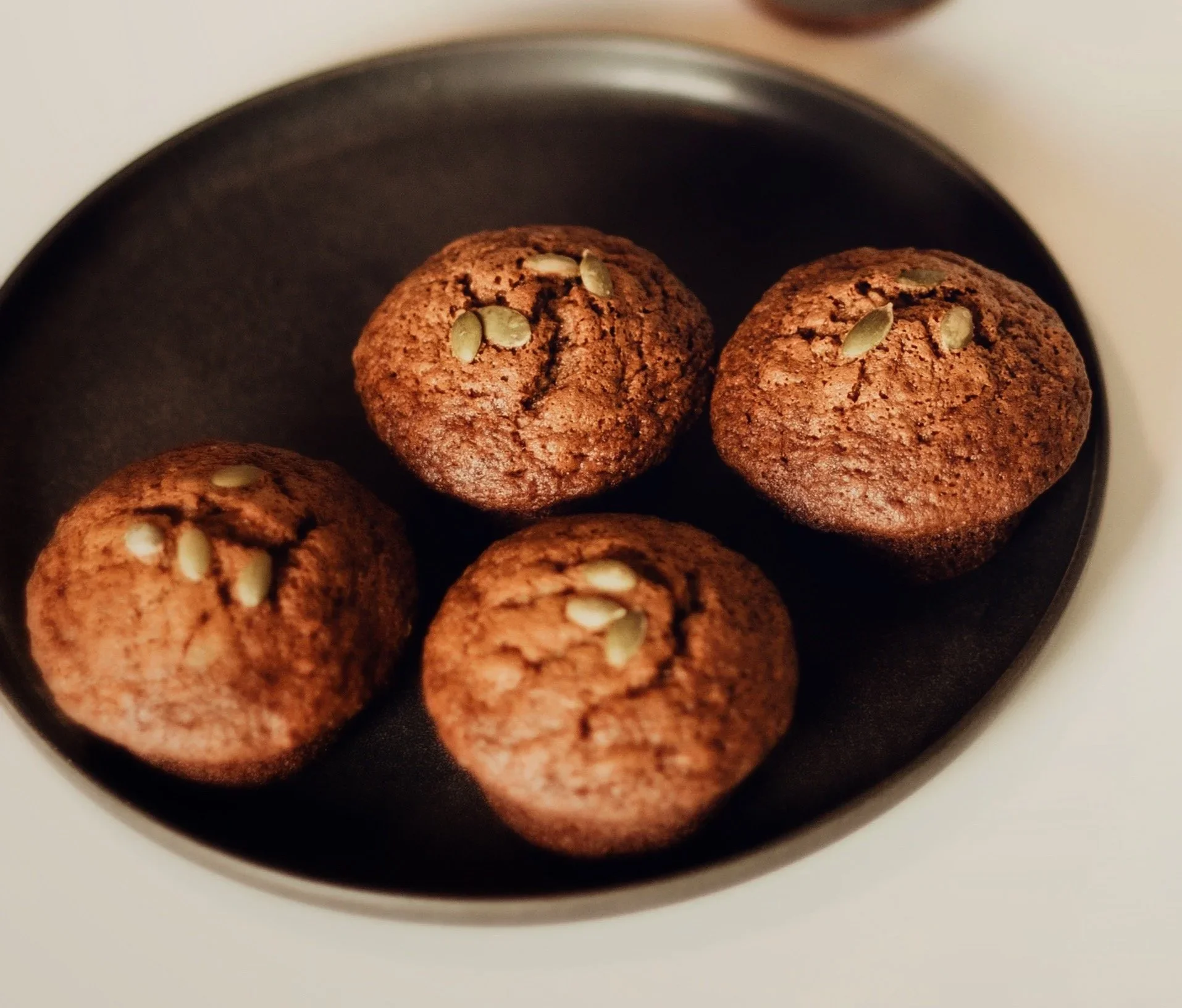 Close-up of chocolate cookies with pumpkin seeds on top, inside a black bowl on a white surface.