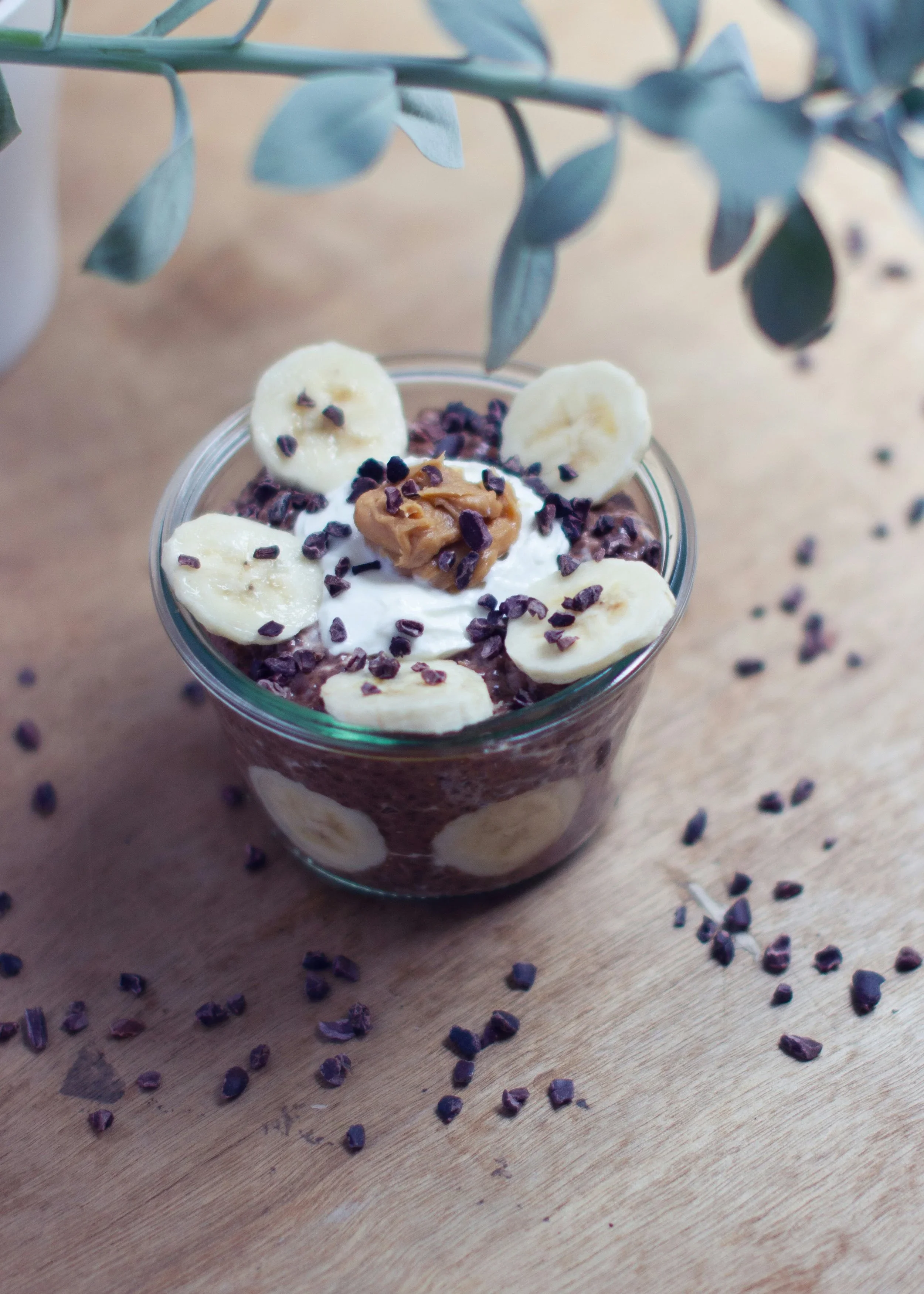 A clear glass filled with chocolate pudding topped with banana slices, a dollop of peanut butter, chocolate sprinkles, and surrounded by scattered chocolate sprinkles on a wooden surface.