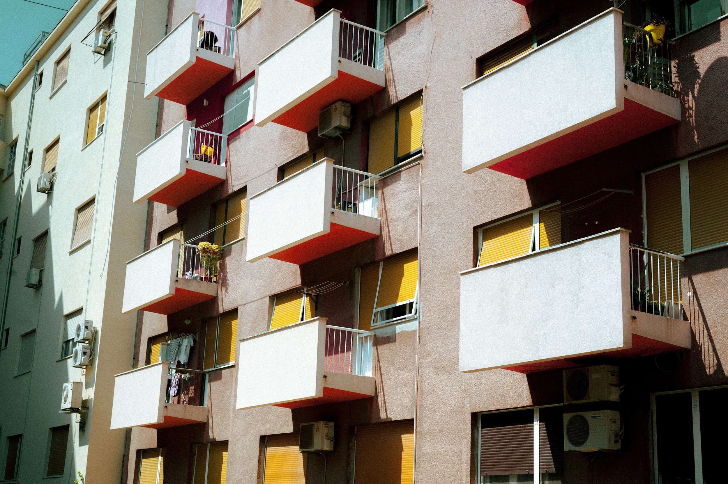 Multiple apartment balconies on a building, with some plants, laundry, and yellow window blinds, casting shadows on the wall.