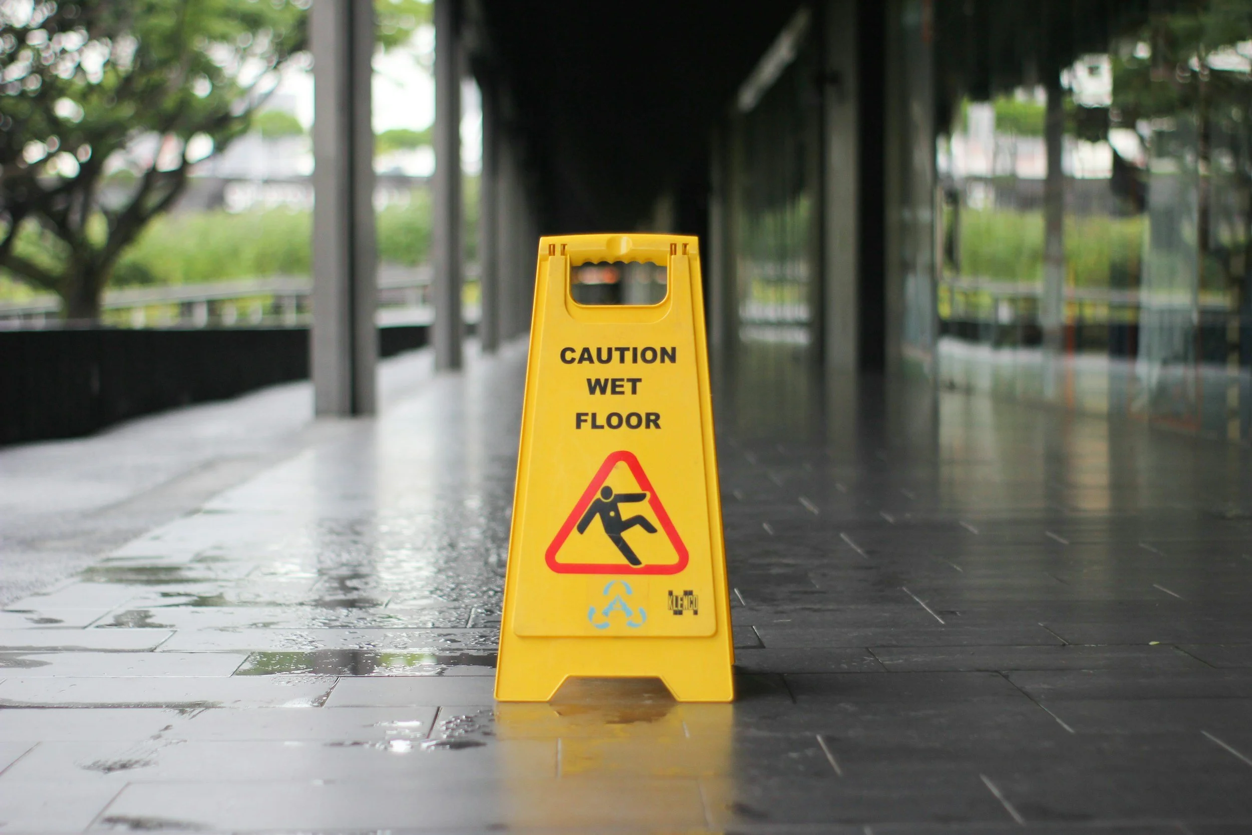 Yellow wet floor caution sign with a person slipping symbol, placed on a wet tiled floor in an indoor or covered area.
