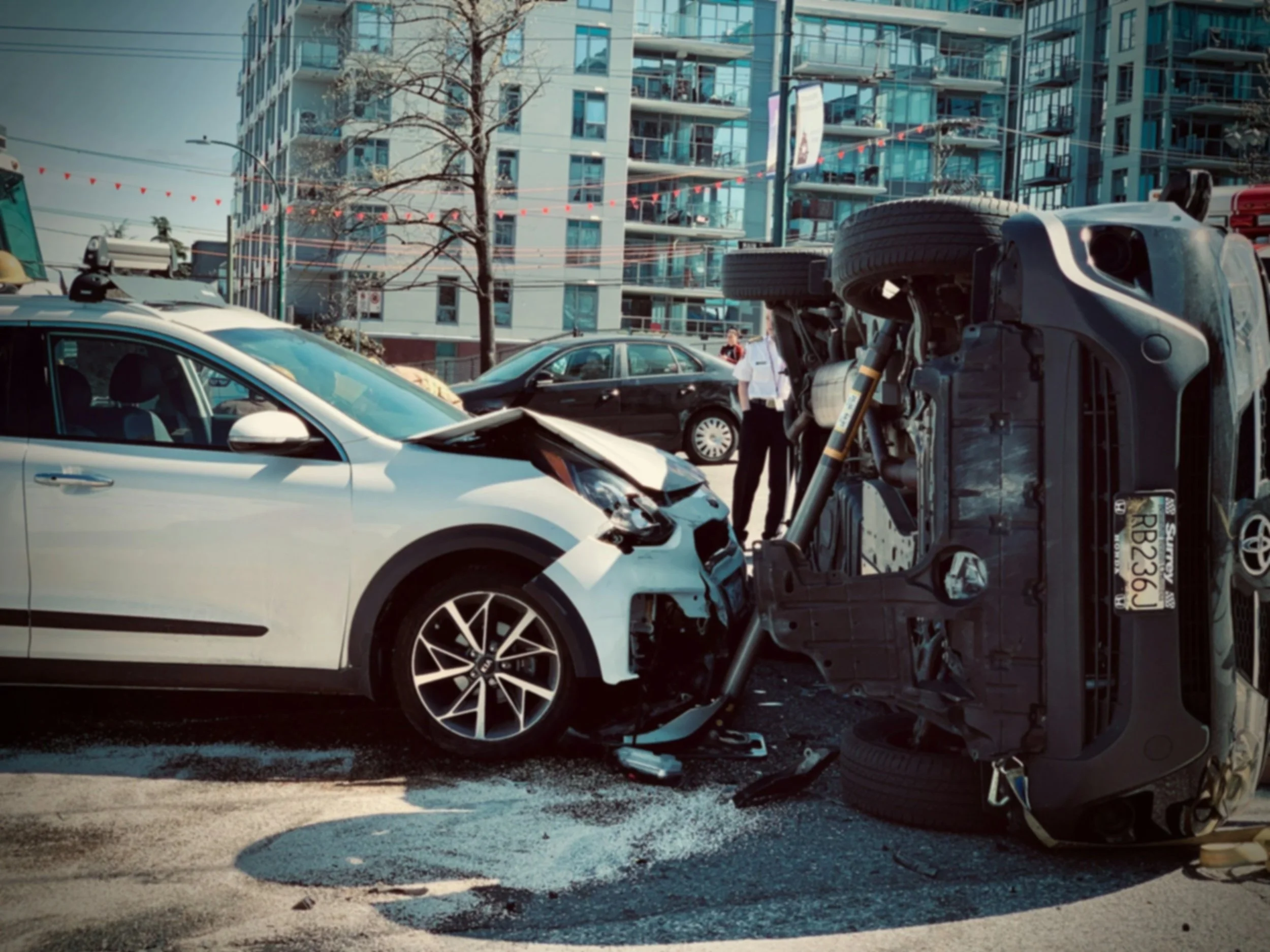 A car accident with a white car with front-end damage and a overturned vehicle in an urban area with high-rise buildings in the background.