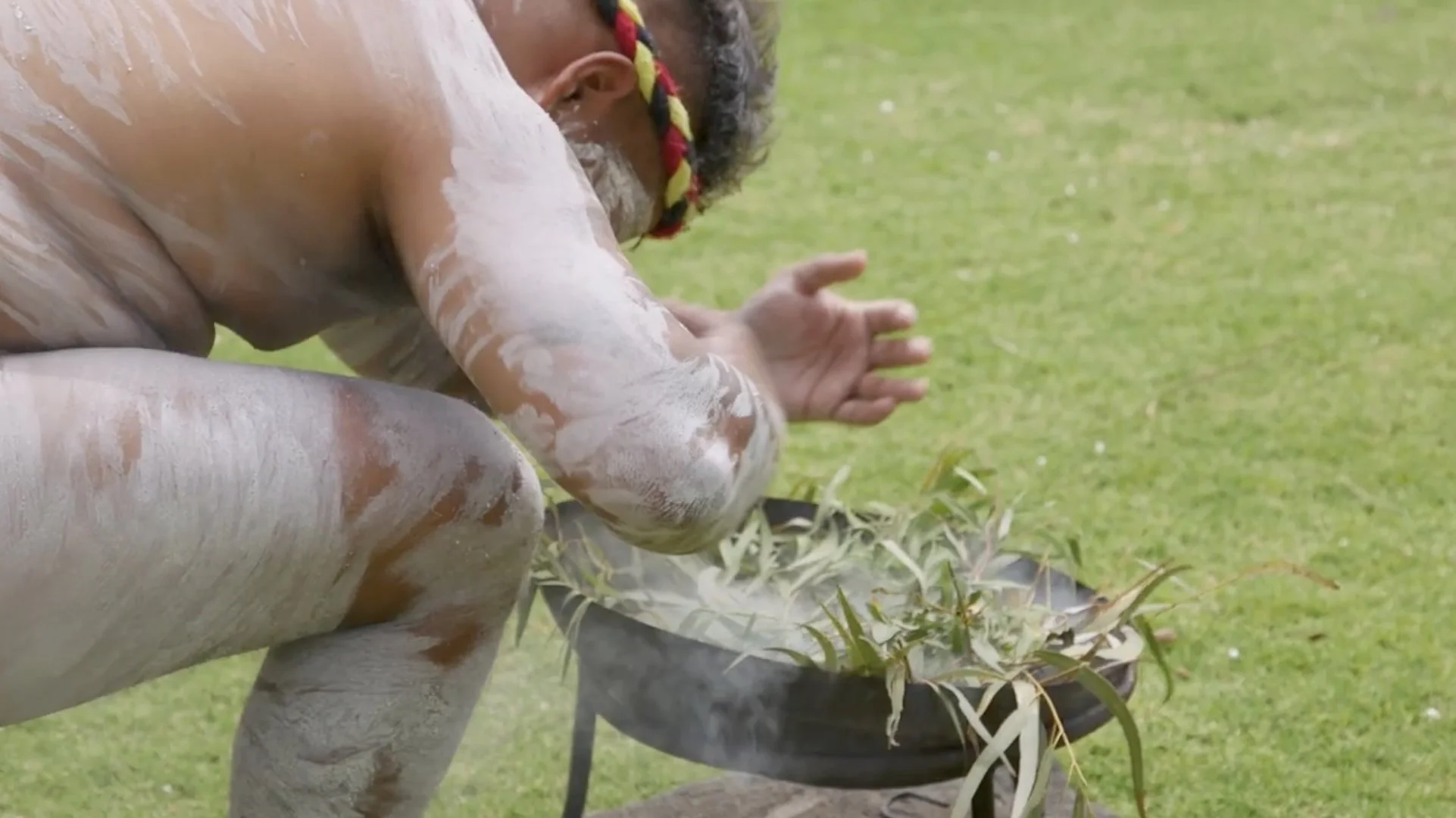An Aboriginal with traditional body paint, wearing a braided headband, crouching during a traditional smoking ceremony.