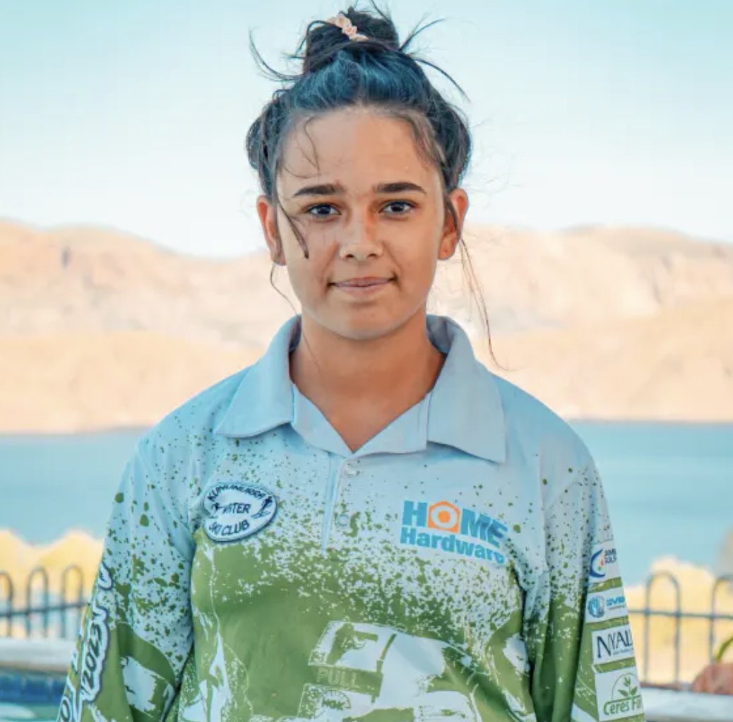 Young girl with a bun hairstyle, standing outdoors near water and mountains, wearing a blue and green sports jersey with sponsor logos.