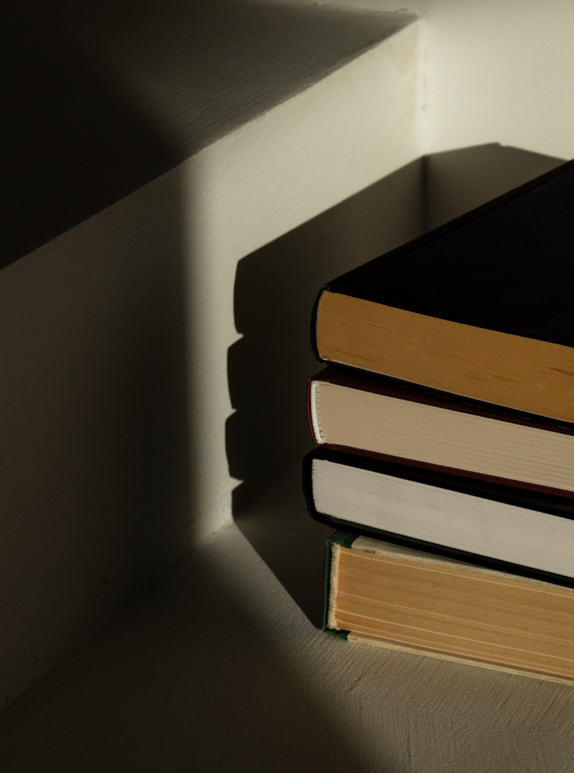 A stack of four books with shadows cast on a white wall in the corner of a room.