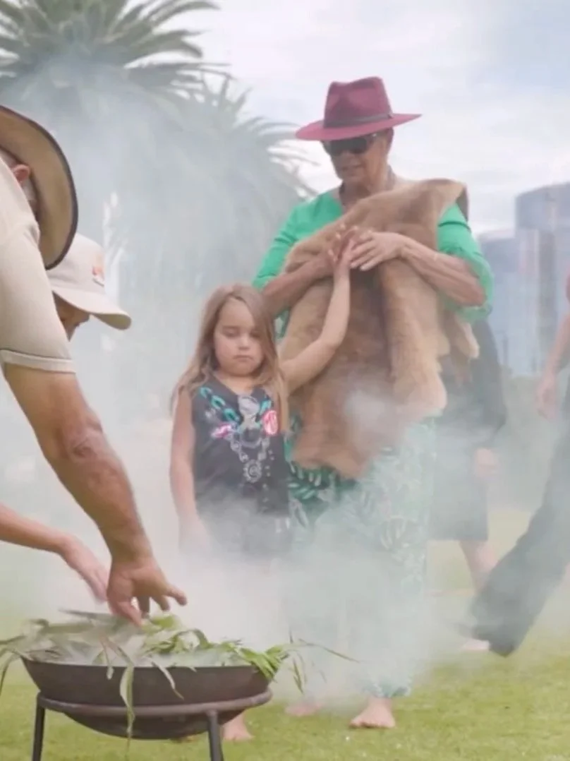 A group of people outdoors, surrounding an Aboriginal smoking ceremony. 