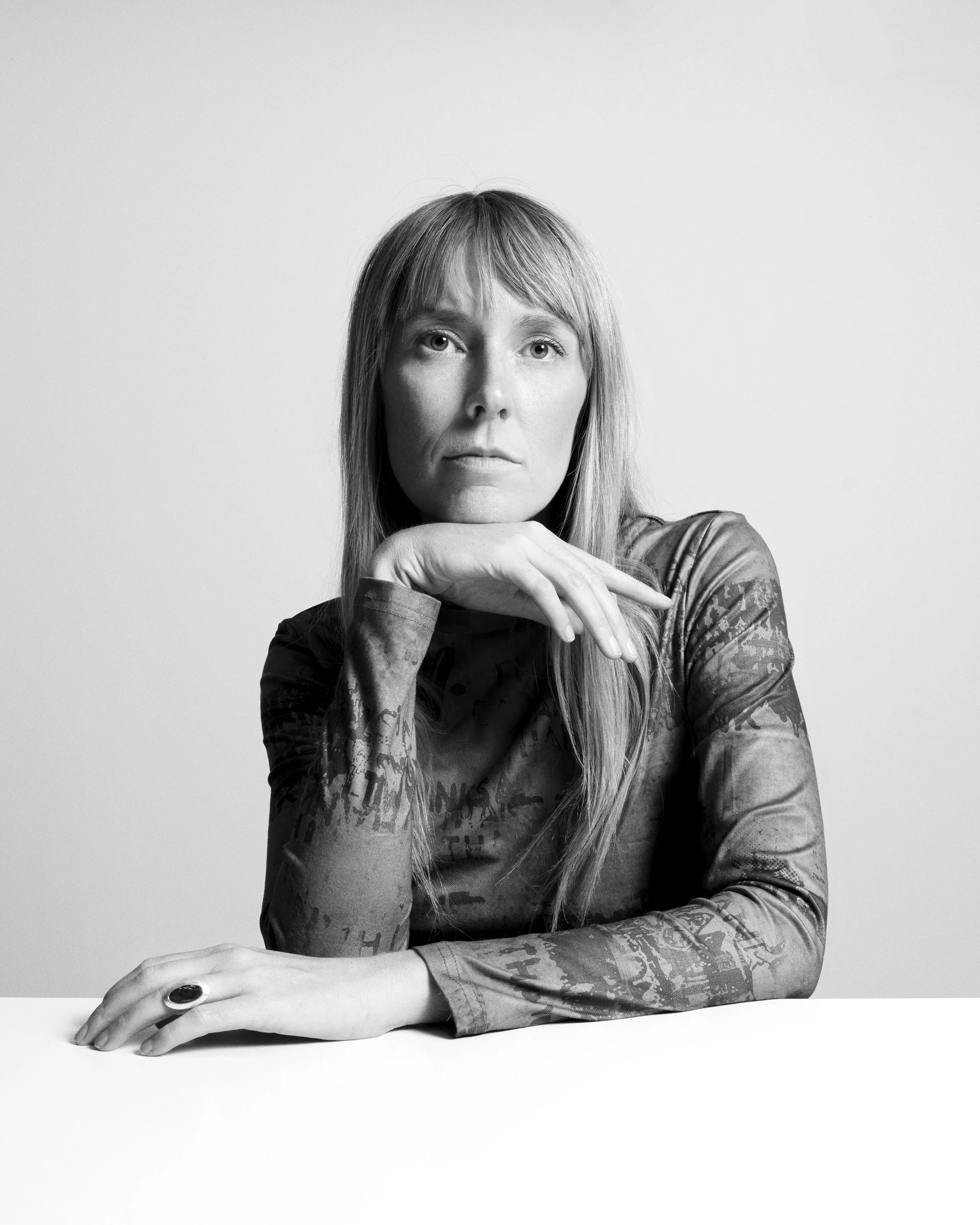 Black and white portrait of Clare Reid, resting her hand on her chin, wearing a patterned long-sleeve shirt.