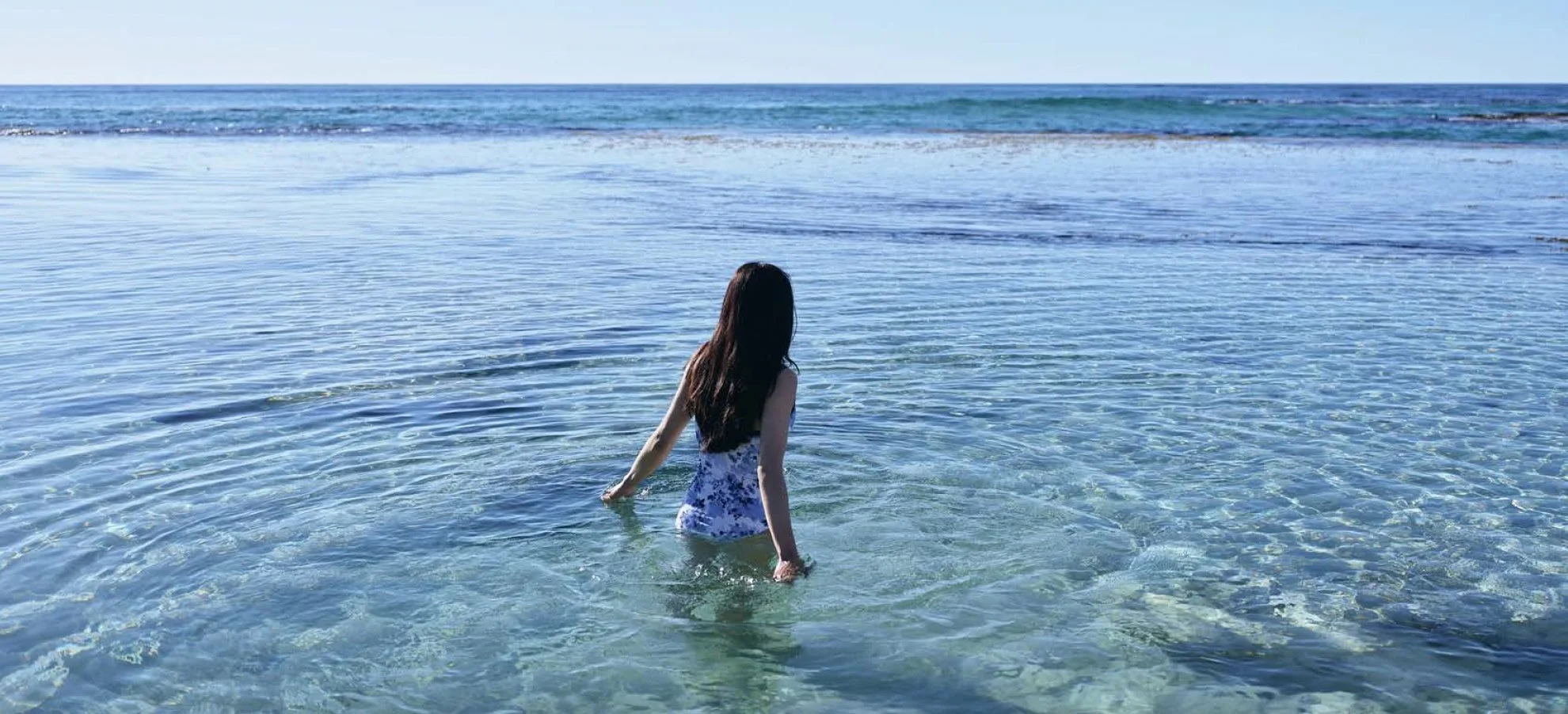 A woman with long brown hair wearing a floral dress stands knee-deep in clear ocean water, looking towards the horizon under a blue sky.