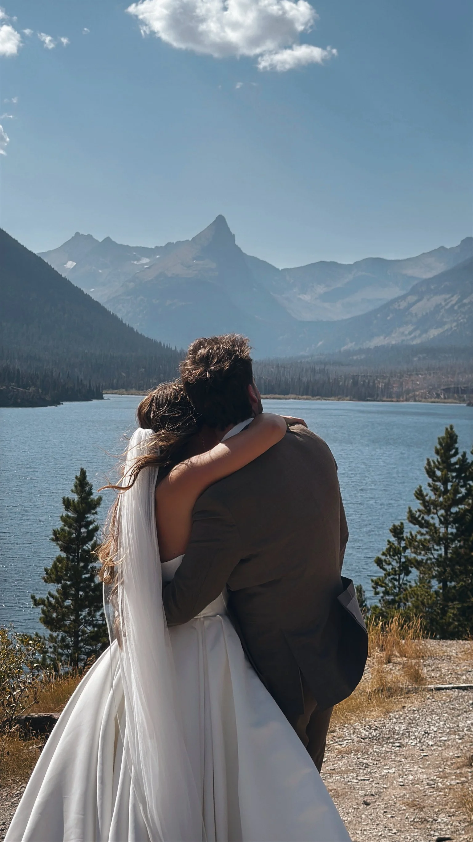 A couple hugging outdoors with a mountain lake and mountains in the background.