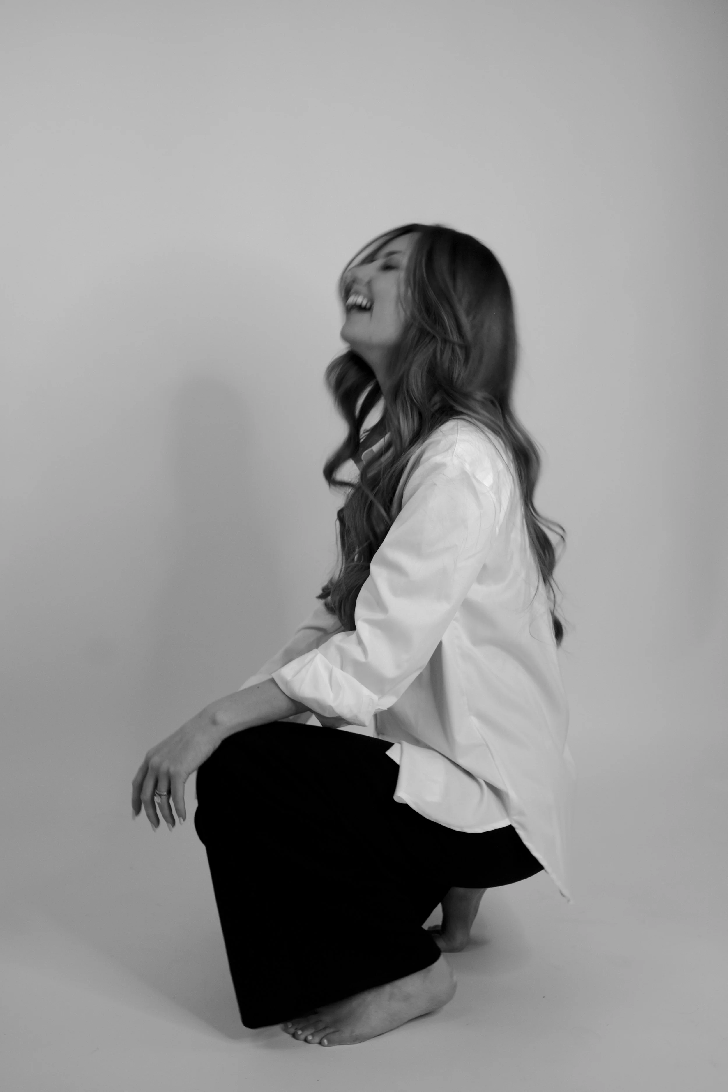 A woman with long hair laughing, sitting on her knees with her hand on her knee and her head tilted back, wearing a white shirt and black pants, against a plain background.