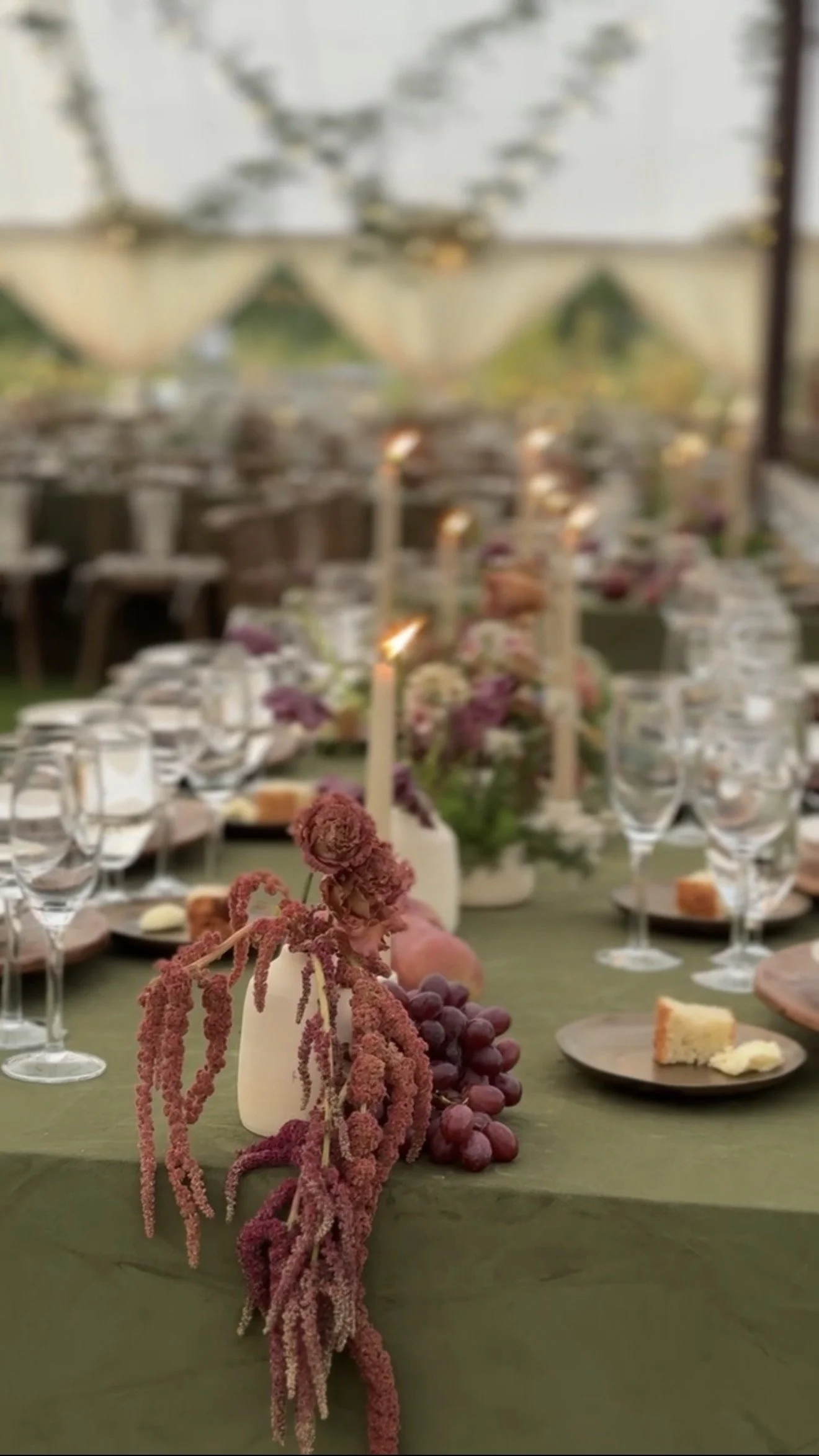 Decorative tablescape with pink and purple flowers, grapes, candles, and cake slices on a green tablecloth at a wedding or celebration event.